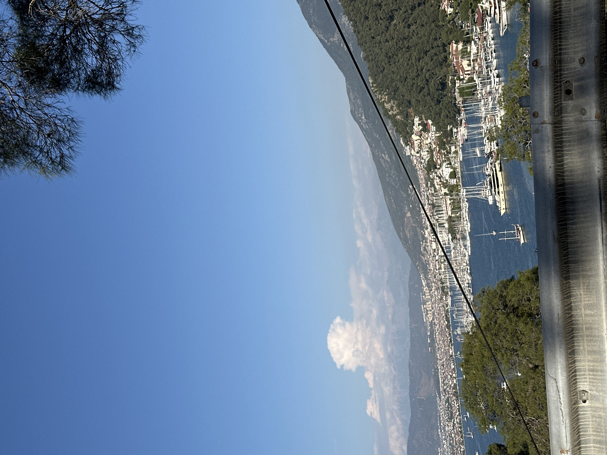 The image displays a panoramic view of a coastal town and harbor, identified as Fethiye, Turkey, under a clear, bright blue sky with sparse white clouds. The perspective is from an elevated viewpoint, looking down upon the expansive scene.
In the midground, a tranquil blue body of water, likely a bay or marina, is densely populated with numerous boats, predominantly white sailboats and yachts with tall masts. Along the curving shoreline, a bustling town extends, characterized by a cluster of white or light-colored buildings interspersed with greenery. Behind the town, a prominent mountain range covered in dense green forests rises, dominating the background and fading into a light haze further in the distance.
In the immediate foreground, the upper left corner features the dark, intricate branches of a tree. On the right side, a gray, metallic structure, possibly a railing or support, is visible with bolts and a textured surface. A thin, dark, diagonal line, resembling a cable or wire, stretches across the frame from the upper left towards the lower right.
The scene suggests a clear and sunny daytime, likely morning or early afternoon, with excellent visibility. No visible text is present in the image.