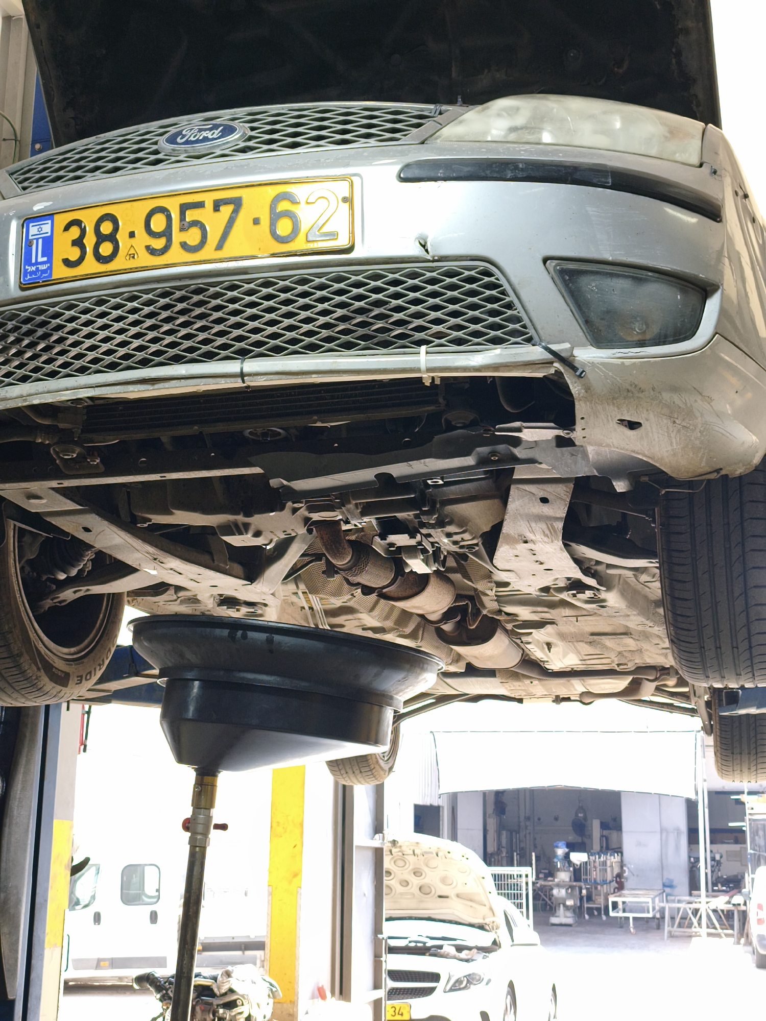 The image shows the underside of a silver Ford sedan being worked on in an auto repair shop. The car is elevated on a lift, and an oil drain pan is positioned beneath the engine. A license plate with the number "38-957-62" is visible on the front of the car. The background shows the interior of the workshop, with tools and equipment present. The overall scene suggests a routine car maintenance operation.