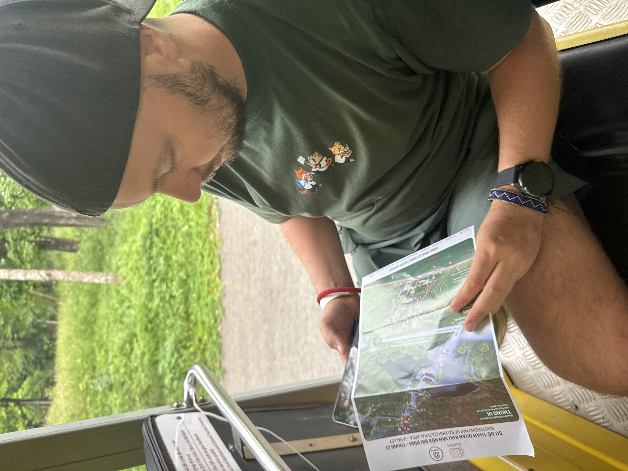 In the city of Tây Hoa Lư Ward, Vietnam, a person wearing a dark green t-shirt with a graphic of four cats and a dark baseball cap is seated inside a vehicle. The person is holding and looking at a map labeled "SỞ ĐỒ THAM QUAN KHU VĂN HOÁ BÁI ĐÍNH - THUNG LŨI CULTURAL AREA - UI VALLEY". The map appears to detail a tourist area with various points of interest. The person has a red bracelet on their left wrist and a smartwatch with a dark strap on their right wrist. The background shows a lush green environment with trees and foliage, suggesting an outdoor setting. The vehicle interior is visible with a yellow seat and metal flooring.