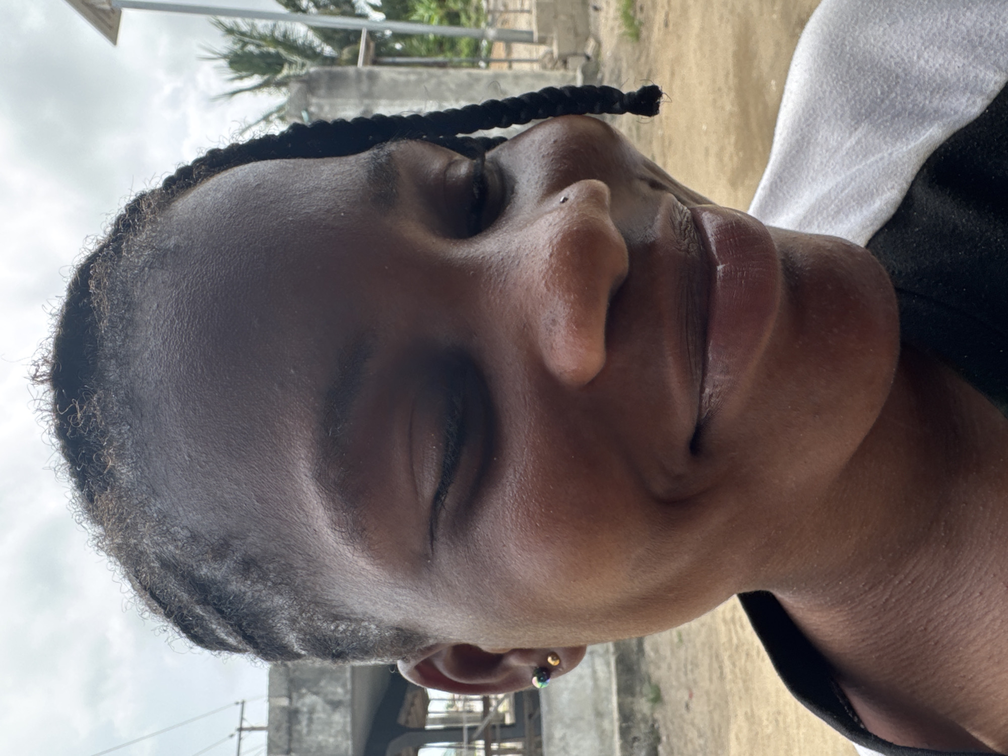 A close-up portrait shows a woman with dark skin, her eyes closed. Her hair is styled in braids. She has a nose piercing in one nostril. She is wearing earrings. The backdrop includes elements of a construction site with scaffolding and other buildings. The sky is overcast. Palm trees are visible, suggesting a tropical environment. Due to the prompt's mention of Badagry, Nigeria, the location is presumed to be in that city.