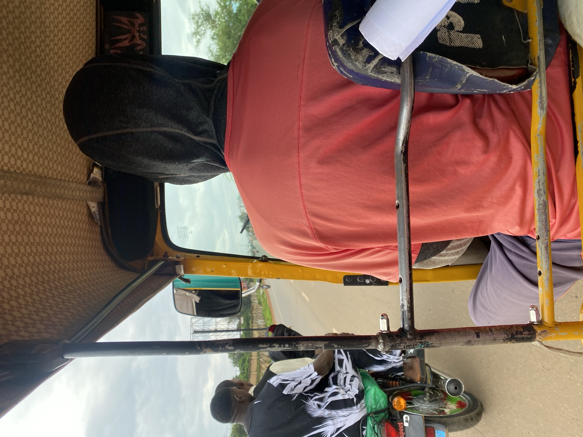 The image, taken from the inside of a yellow auto-rickshaw, shows a driver wearing a dark hoodie and a coral-colored shirt, with his back to the camera. To the left, two other individuals are visible in the auto-rickshaw. One, wearing a black and white patterned shirt with palm tree designs, is seated in the front passenger area. Another individual is partially visible behind him. A motorcycle is seen beside the auto-rickshaw, with a green cover over its seat and a visible license plate with the word "Respect". The auto-rickshaw is traveling on a paved road, with trees and a cloudy sky visible in the background. The overall environment suggests an outdoor setting in what appears to be Rafin Par, Nigeria.