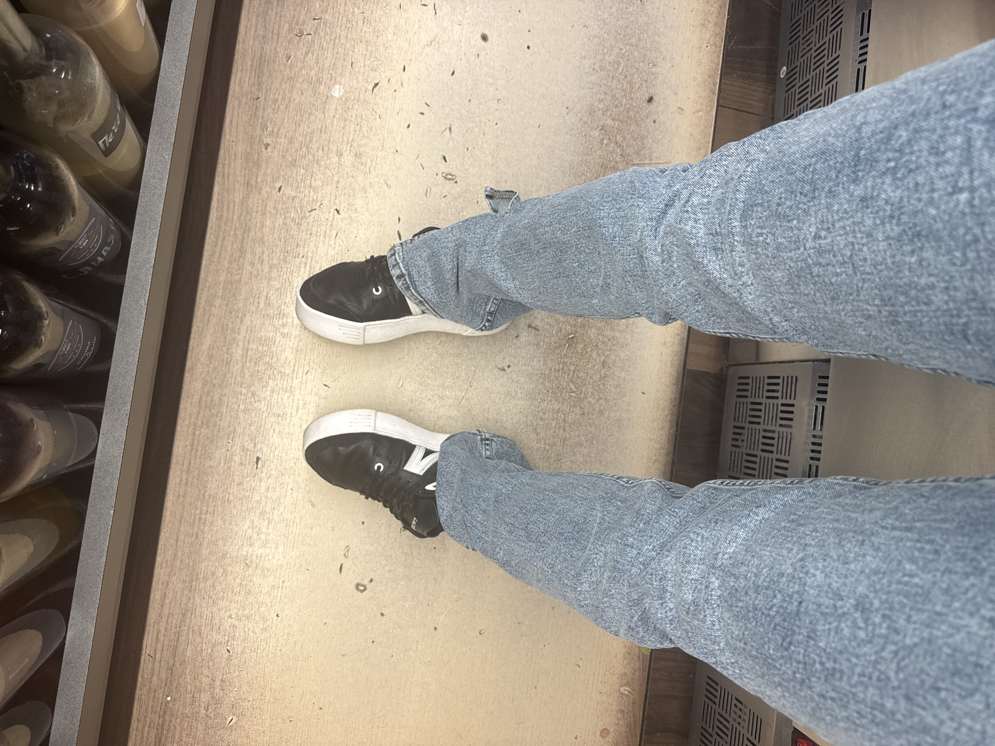The image shows a person standing indoors. The person is wearing blue jeans and black sneakers with white soles. The sneakers have a white "N" logo. To the left of the person, there is a rack filled with wine bottles. To the right of the person, there is a metal grate. The floor is beige and has dirt spots. There is no evidence of the location being Moscow, Russia.