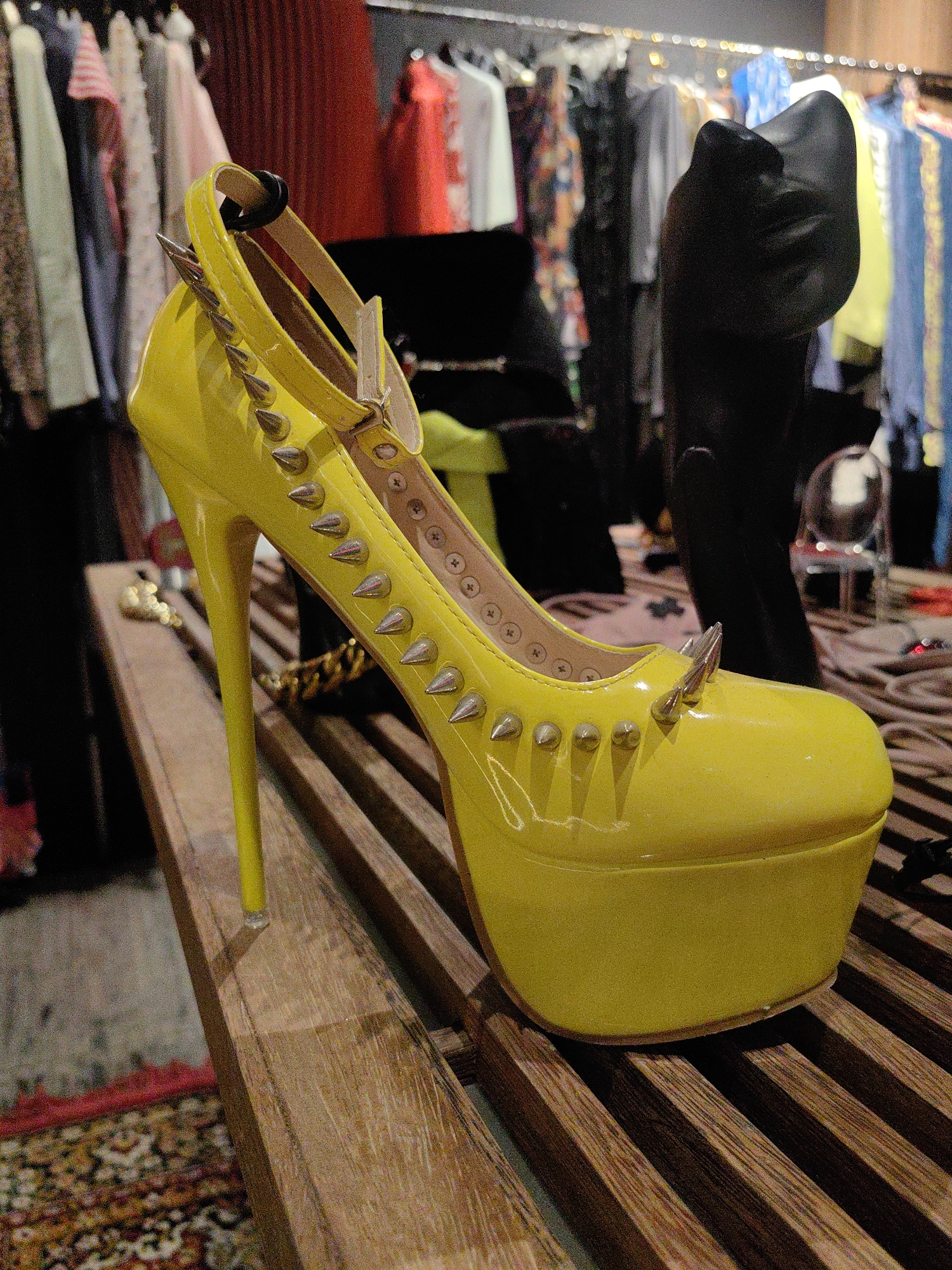A bright yellow platform high-heeled shoe adorned with silver spikes is displayed on a wooden slatted surface. The shoe features a thin stiletto heel and a strap that buckles around the ankle. The platform is substantial, and the spikes are arranged along the edges of the shoe, including the toe and the sides. In the background, racks of clothing are visible, suggesting a retail or display setting. The lighting is artificial, and the overall impression is of a fashion-forward or alternative style of footwear. No people are visible in the image. The location context provided is Baan Mae Nam, Thailand, which does not appear to be directly reflected in the visual cues of the image itself.