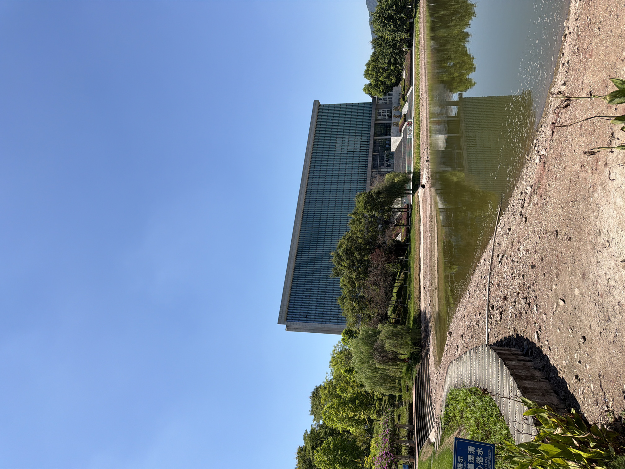 The image shows a modern building with a glass facade reflected in a calm lake. In the foreground, a curved pathway made of wooden planks runs along the edge of the lake, bordered by gravel and lush green vegetation. A blue sign with Chinese characters is posted on the left side of the pathway. Behind the pathway, a grassy slope rises towards the building, dotted with trees of various sizes and lush foliage. The building itself is a rectangular structure with a long, horizontal design, featuring numerous glass windows that reflect the clear blue sky. The entrance to the building is accessed via a wide set of stairs leading up to the main doors, with signage above. The entire scene is bathed in bright sunlight, suggesting a clear day. This appears to be the library building of Chongqing University of Humanities and Sciences in Jiangjin, China.