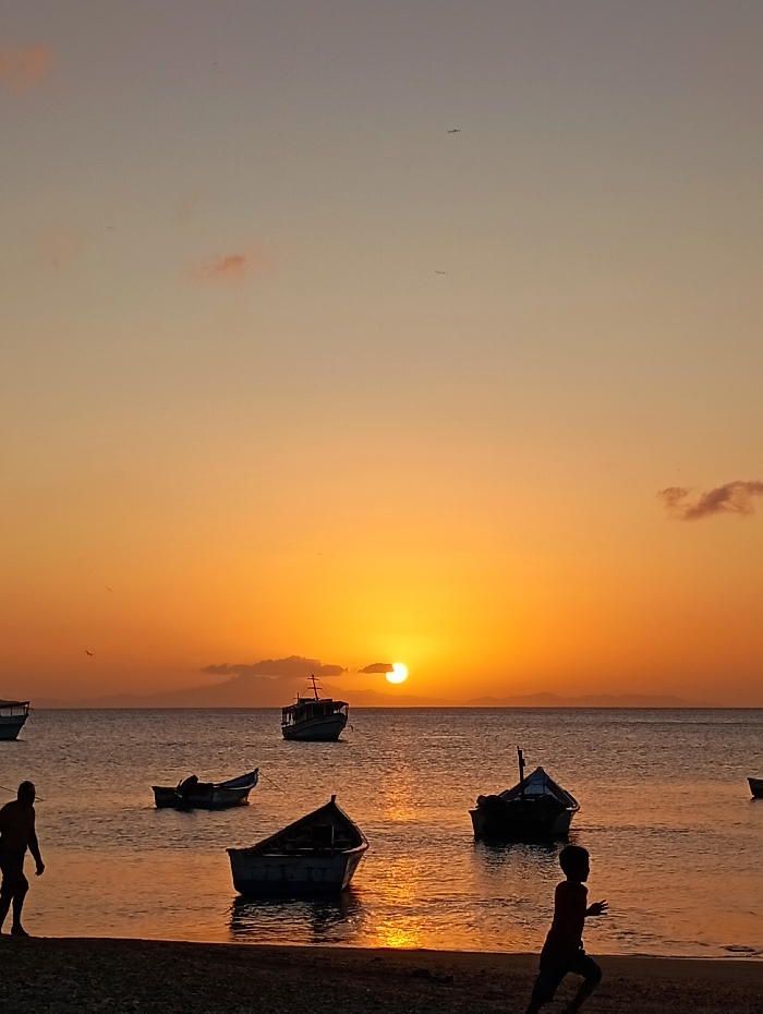 Golden sunlight illuminates a tranquil beach scene at sunset. Several small boats float gently in the calm, reflective water. Two silhouetted figures stand on the shore, one appearing to walk while the other runs. The sky is a gradient of orange and yellow hues, with a few wispy clouds near the horizon. The sun, partially obscured by distant land, casts a warm glow over the entire scene.