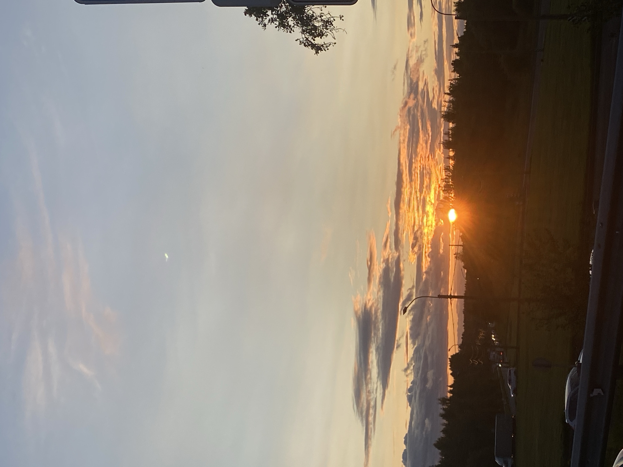 The image depicts a sunset over Peterhof, Russia, with the sun setting behind a dense line of trees that form the horizon. The sky is a gradient of warm colors, transitioning from a pale yellow near the horizon to a soft blue at the top, with scattered clouds tinged with orange and pink. A crescent moon is visible in the upper portion of the sky. In the foreground, a road is visible with several vehicles, including cars and trucks, driving on it. Streetlights are also present along the road, casting a faint glow. The scene appears to be a typical evening commute in a suburban or semi-rural area, with nature and urban elements coexisting. The overall mood is serene and peaceful, characteristic of a sunset.