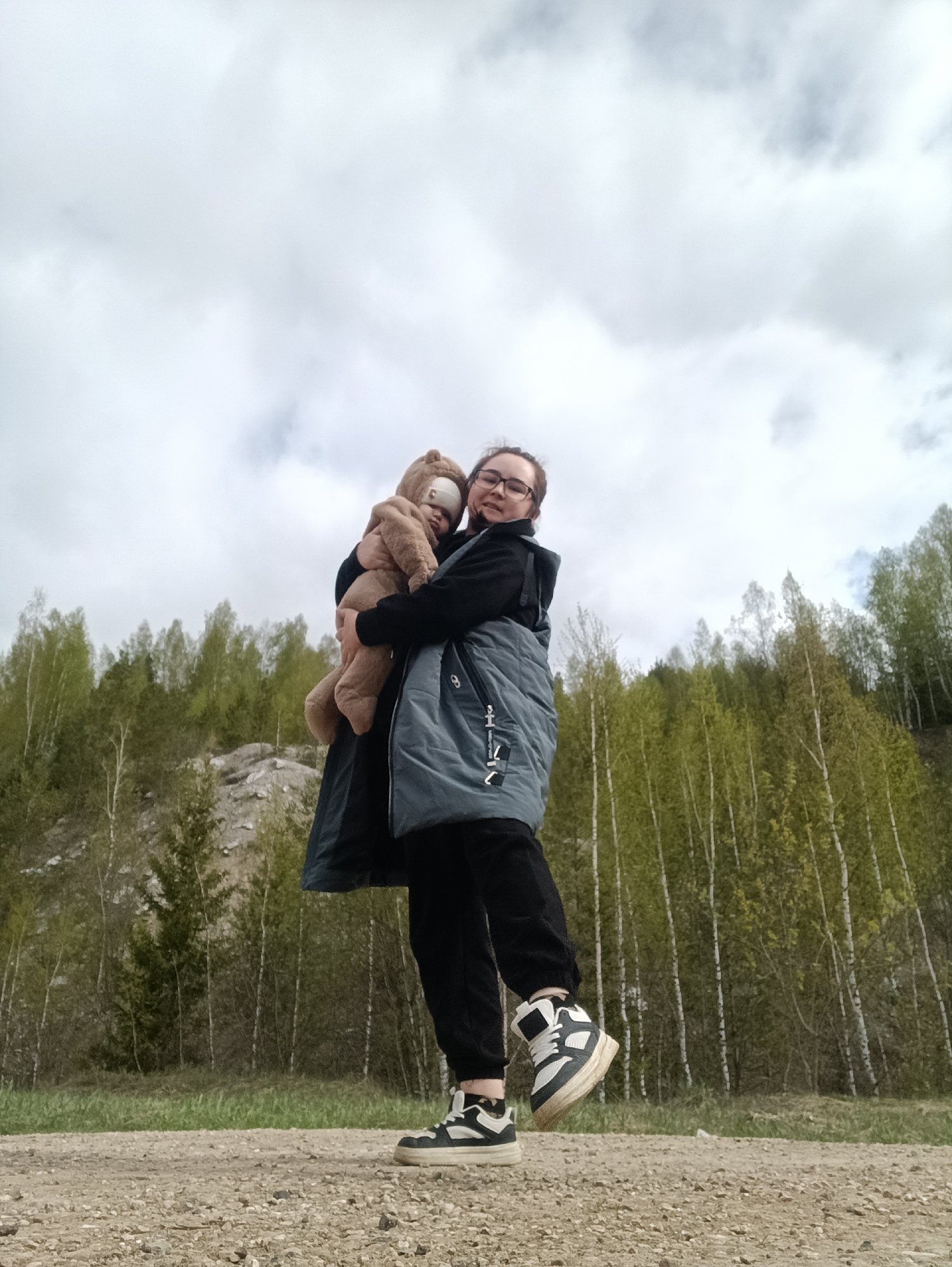 A woman in glasses, a black coat, and a grey vest holds a baby in a tan hooded jumpsuit. They are standing on a gravel path, with a background of trees on a hillside and a cloudy sky. The location is Посад, Russia.