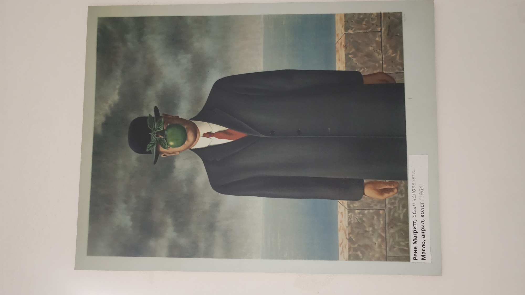 The image shows a reproduction of René Magritte's painting "The Son of Man" (1964). The subject is a man in a dark suit and bowler hat standing near what appears to be a low wall with the sea and sky in the background. An apple obscures his face. A label on the image indicates the painting's name, artist, materials used (oil, acrylic on canvas), and date (1964). The image does not depict any specific location in Moscow, Russia.