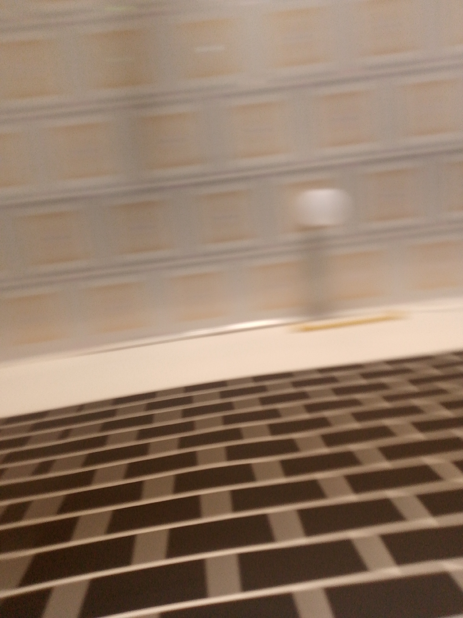 The image displays a blurred interior scene, likely a hallway or room, with a tiled floor in the foreground and a wall with rectangular patterns and a diffused light source in the background. The floor is composed of dark and light rectangular tiles arranged in a brick-like pattern. The wall above the floor has horizontal bands of lighter material with darker rectangular insets, suggesting a decorative paneling or wallpaper. A blurred circular light or fixture is visible on the upper portion of the wall. There is no indication of the specific location within Jauro Yinu, Nigeria, nor any people or specific actions taking place. The image's blurriness suggests movement during capture or a focus issue.