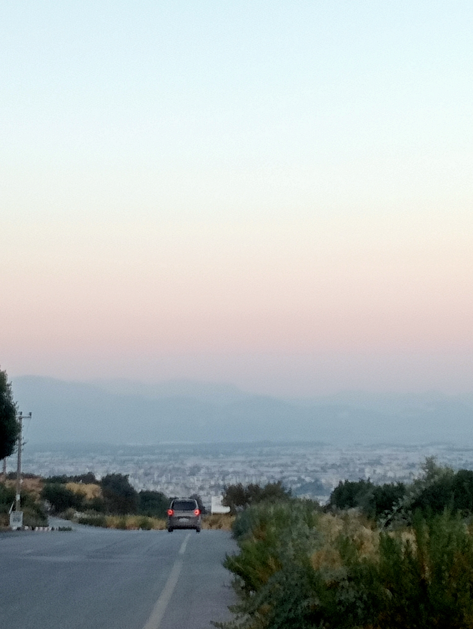 The image depicts a landscape scene with a road in the foreground. A silver car is driving away from the viewer on the asphalt road. The road has a white line marking on the right side. On the left side of the road, there is a utility pole. The background shows a city or town spread out in the distance, with a hazy atmosphere. The sky transitions from a pale blue at the top to a soft pinkish hue towards the horizon, suggesting it is either dawn or dusk. The terrain in the middle ground is hilly and covered with vegetation, including trees and bushes. The overall impression is of a serene, open landscape with a distant urban settlement.