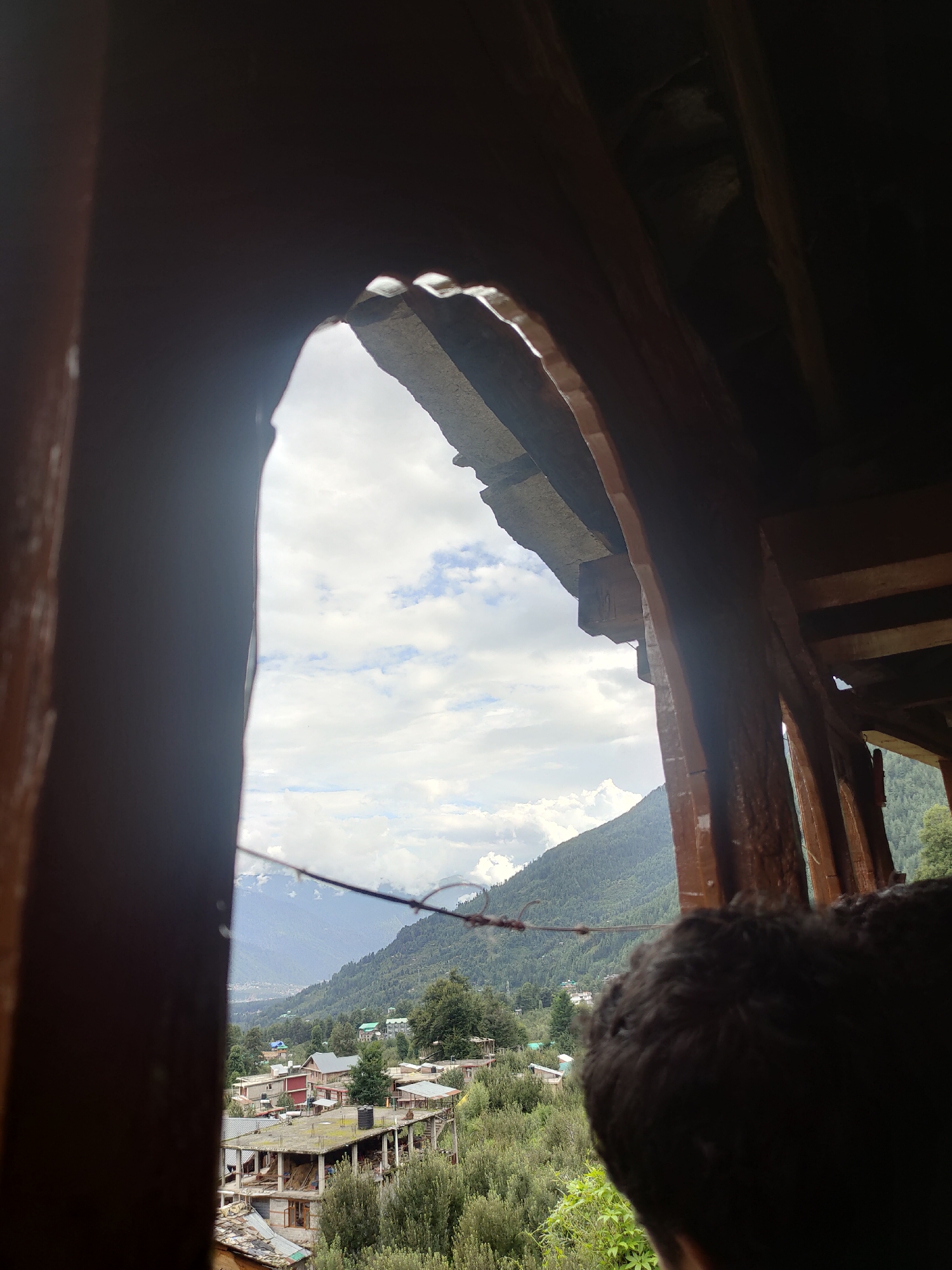 A person's dark, curly hair fills the lower right foreground of the image, obscuring the view to the right. The image is taken from within a wooden structure, looking out through an arched opening. The opening frames a view of a village nestled in a valley, with buildings of various sizes and materials. A mountainous landscape, covered in dense green trees, rises in the background, meeting a cloudy sky. In the distance, more mountains are visible, fading into a hazy blue. The village appears to be located in Burwa, India, given the architectural style and the lush, hilly terrain characteristic of the region. The scene suggests a peaceful, rural setting, with the wooden structure providing a rustic frame for the picturesque vista.