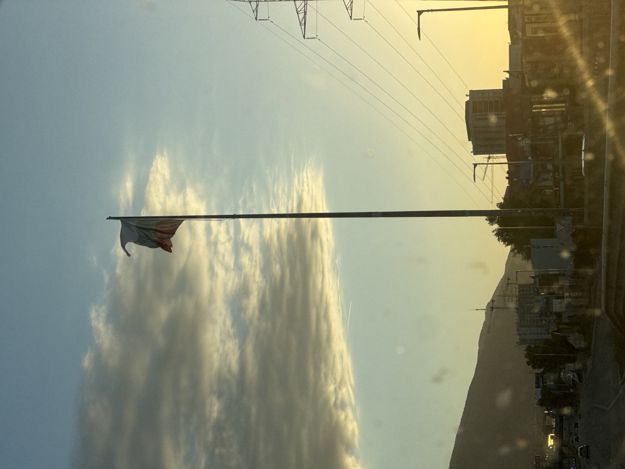 Captured from a low angle, the scene is dominated by a horizontally oriented Bulgarian flag waving in the wind, set against a backdrop of layered clouds and a hazy sky. Above the flag, high-voltage power lines stretch across the upper portion of the image.
In the background, a mountain range defines the horizon, with the urban landscape of what appears to be Sofia, Bulgaria nestled at its base. Buildings and infrastructure, including construction cranes, are visible in the distant cityscape. The overall ambient light suggests either sunrise or sunset, casting a yellowish hue over the scene. The image is slightly blurred, possibly due to the lens of the camera or the conditions of the environment.