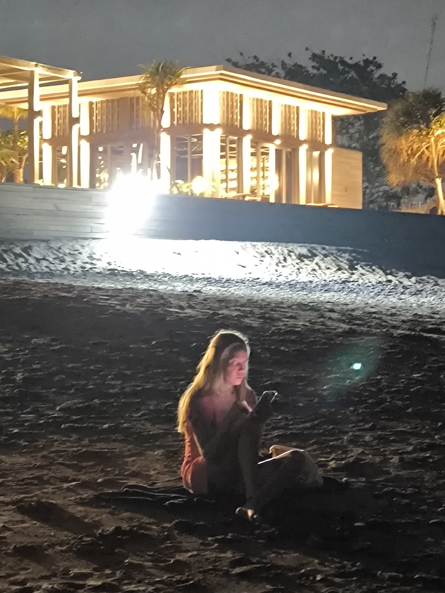 A woman is sitting on a dark beach in Canggu, Indonesia, at night. She is illuminated by the light of her mobile phone, which she is holding in her hands. Behind her is a building with illuminated architectural details and palm trees, indicating a resort or similar establishment. The beach is dark and sandy.