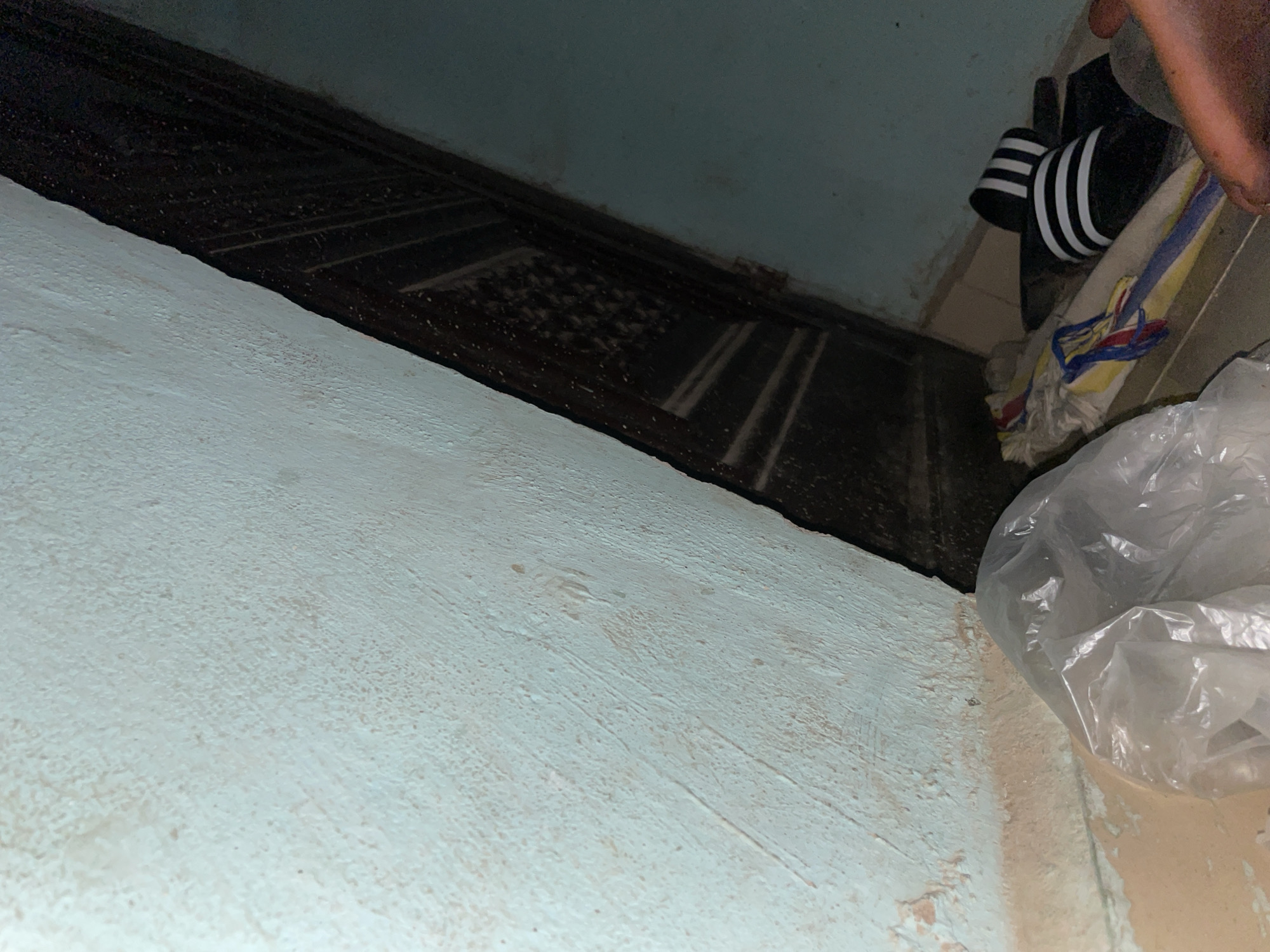 The image depicts an interior corner of a room, likely in Jalingo, Nigeria. A light blue wall, textured with rough plaster, dominates the left side of the frame and extends into the background. A dark, wooden structure, possibly a door or a frame, angles diagonally from the top right to the bottom center, revealing a dark interior with a patterned surface. On the floor in the lower right quadrant, a pair of black and white striped slides rests atop a colorful cloth or mat. In the lower left, a clear plastic bag is visible, partially obscuring the floor which appears to be tiled or made of concrete. A person's right hand and arm are partially visible in the extreme lower right corner, with the hand resting on or near the slides. The lighting suggests an indoor environment, possibly with dim ambient light.