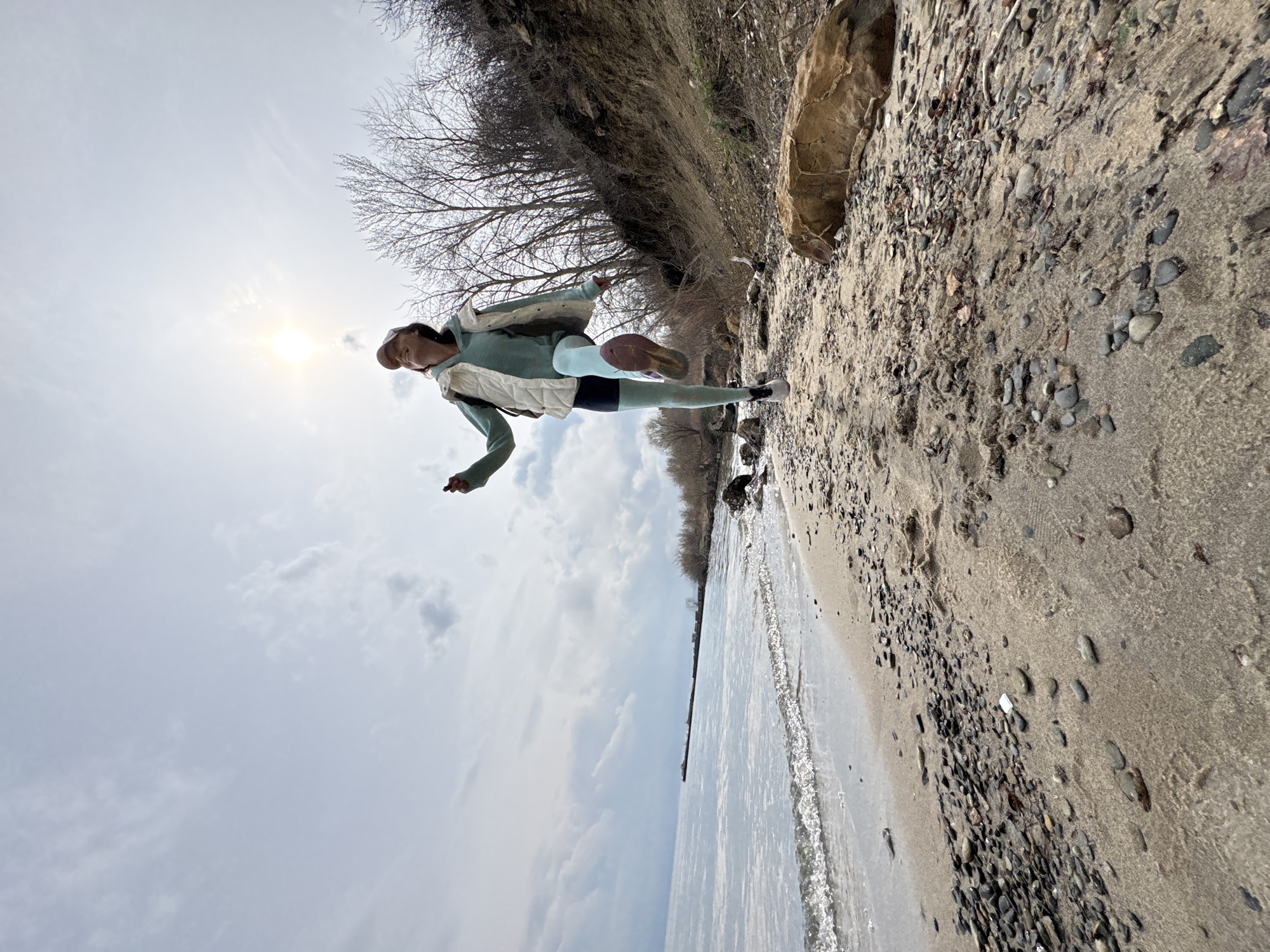 A person in athletic wear is running on a beach next to a body of water under a cloudy sky with the sun visible. The beach is sandy with pebbles, and there is a raised bank or cliff-like edge to the right. Bare trees grow on the edge of the raised bank. The location is Ulyanovsk, Russia.