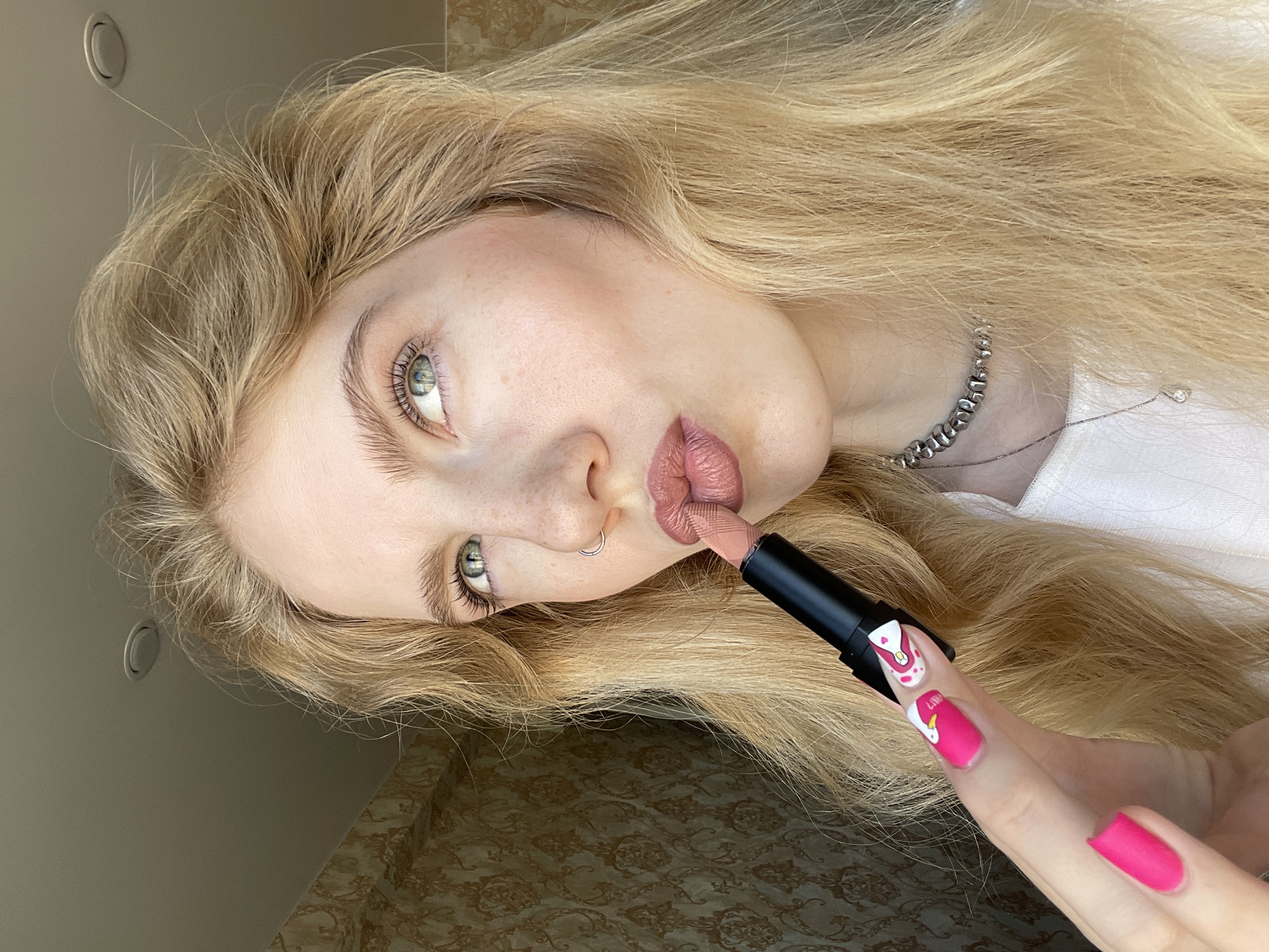 A woman is lying down, applying nude lipstick. She has blonde hair, fair skin, and green eyes. She has a silver nose ring and a beaded silver necklace. She is wearing a white top. Her nails are painted bright pink, with one nail having a cartoon design. The background includes a patterned surface and a ceiling with a circular light fixture. There is no definitive indication of this image being from Izhevsk, Russia.
