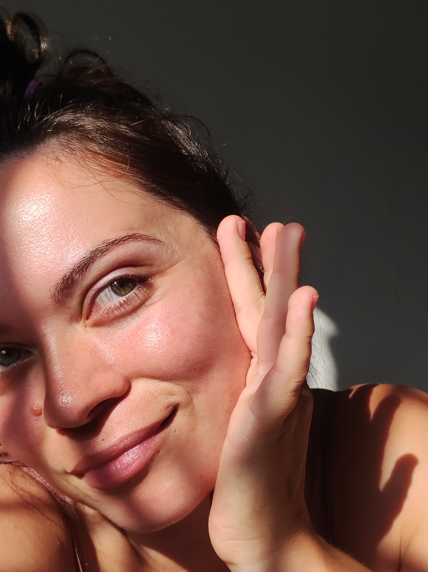 A close-up, low-angle shot captures a woman's face illuminated by dappled sunlight. Her head is tilted to the left, with her right hand delicately placed near her ear, obscuring part of her face. Her eyes, a soft green, are looking towards the viewer, and she has a slight smile on her lips. Her dark hair is pulled back, with a few stray strands framing her face. The background is a plain, dark wall, casting shadows from the light. The image does not contain any discernible elements indicating a specific location in Chaikovsky, Russia.