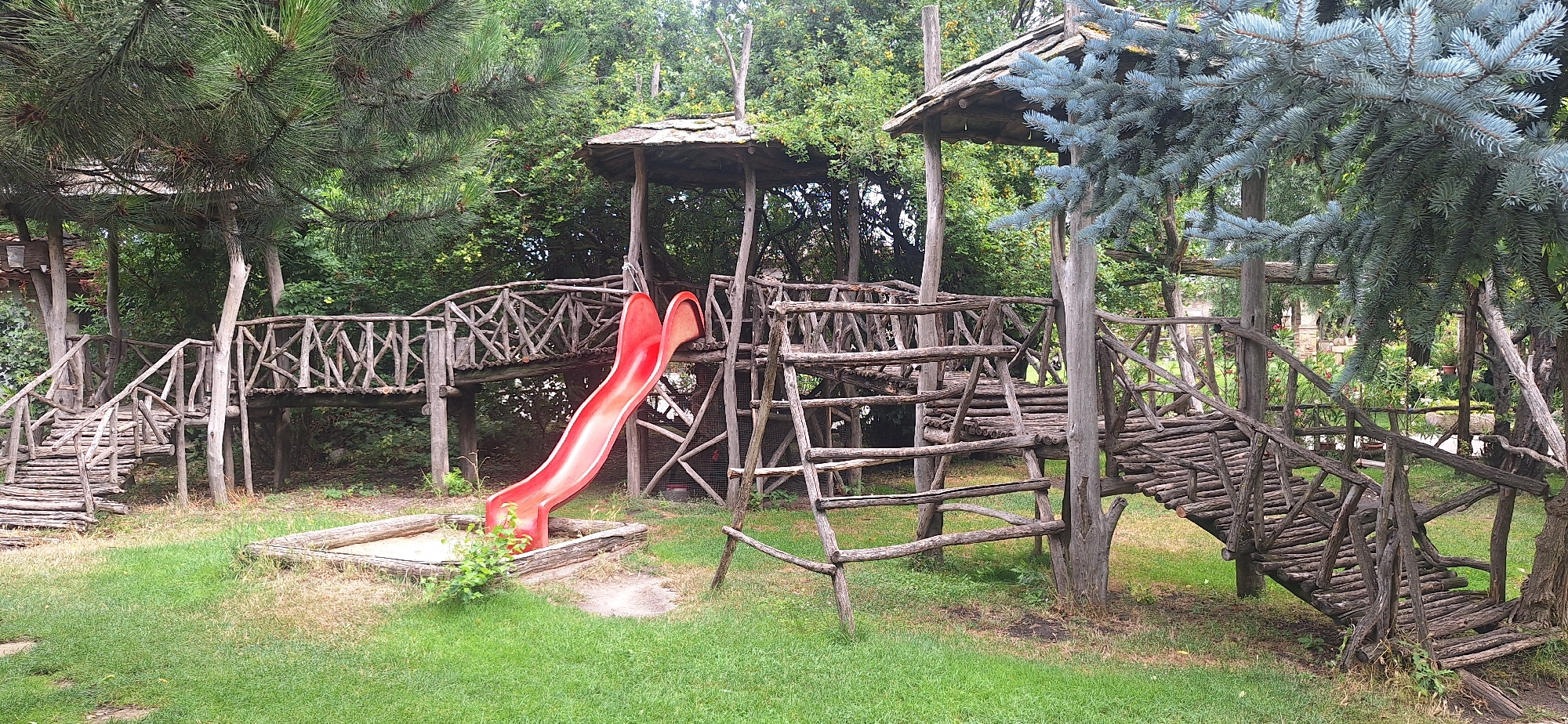 The image depicts a rustic, wooden playground set within a green, wooded area. The playground features multiple elevated platforms connected by bridges made of logs and branches. A bright red slide is prominent, descending from one of the platforms into a sandy pit surrounded by logs. A ladder made of wood is also visible. The surrounding environment is lush with trees and grass. This playground is located in Kalimantsi, Bulgaria. There are no people visible in the image.
