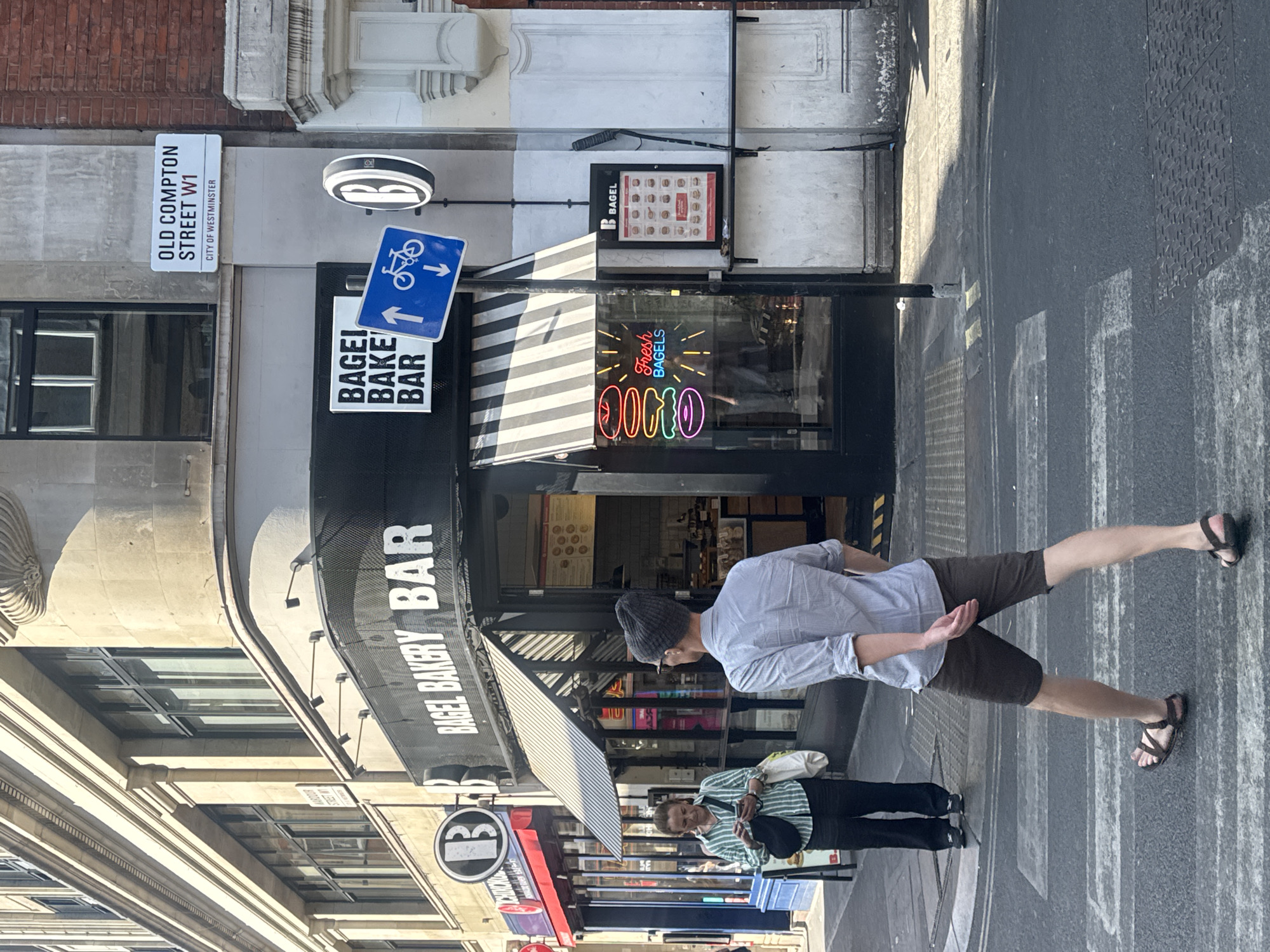 An outdoor, eye-level, medium shot captures a street scene in Westminster, London, United Kingdom, on a sunny day. In the foreground, a person wearing a light blue collared shirt, dark brown shorts, and dark brown sandals walks across a crosswalk. To the left of the crosswalk, a woman wearing a green and white striped long-sleeved shirt and black pants stands on the sidewalk, looking at her phone. She carries a black shoulder bag and a white tote bag. Behind her, a blue awning covers a shopfront.
The central focus of the image is a storefront with a black awning and the words "BAGEL BAKERY BAR" in large white lettering. Below the awning, a neon sign displays "Fresh Bagels" with colorful illustrations of bagels. To the right of the storefront is a white sign with a black border and a black "B" in a circle, indicating "B BAGEL." Adjacent to this sign is a framed menu displaying various bagel options. Above the storefront, a street sign reads "OLD COMPTON STREET W1" and "CITY OF WESTMINSTER." A blue traffic sign with a bicycle symbol is mounted on a pole. The buildings surrounding the scene are made of stone and brick. The street is paved with asphalt, and there are yellow and white road markings for the crosswalk and traffic lanes.