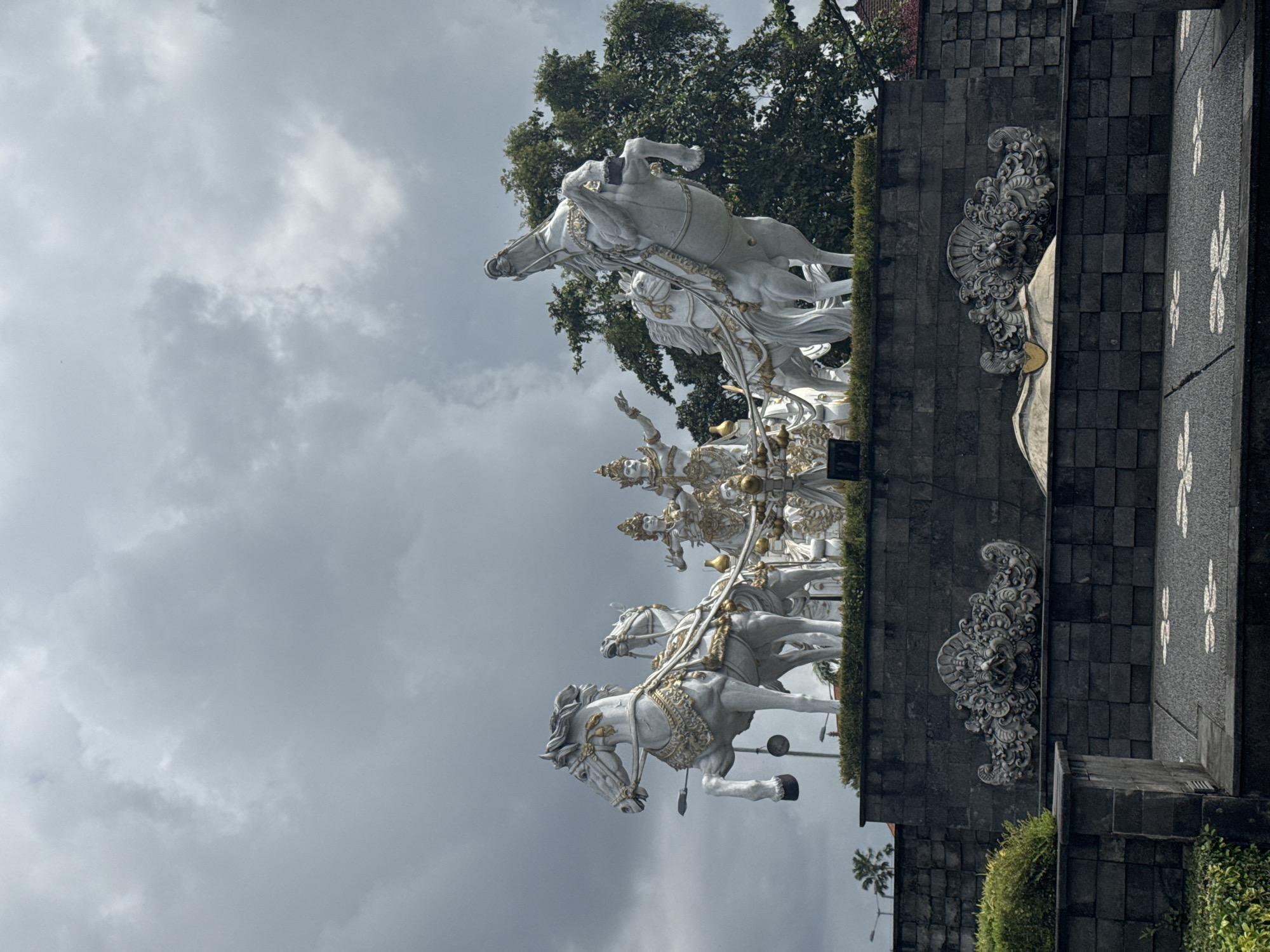 A medium shot shows a large, white statue of a chariot pulled by horses. The statue is on top of a dark grey brick wall with decorative floral carvings. The sky is overcast with grey clouds. The statue appears to be in an urban environment with trees and plants nearby. It’s in Unknown, Unknown.