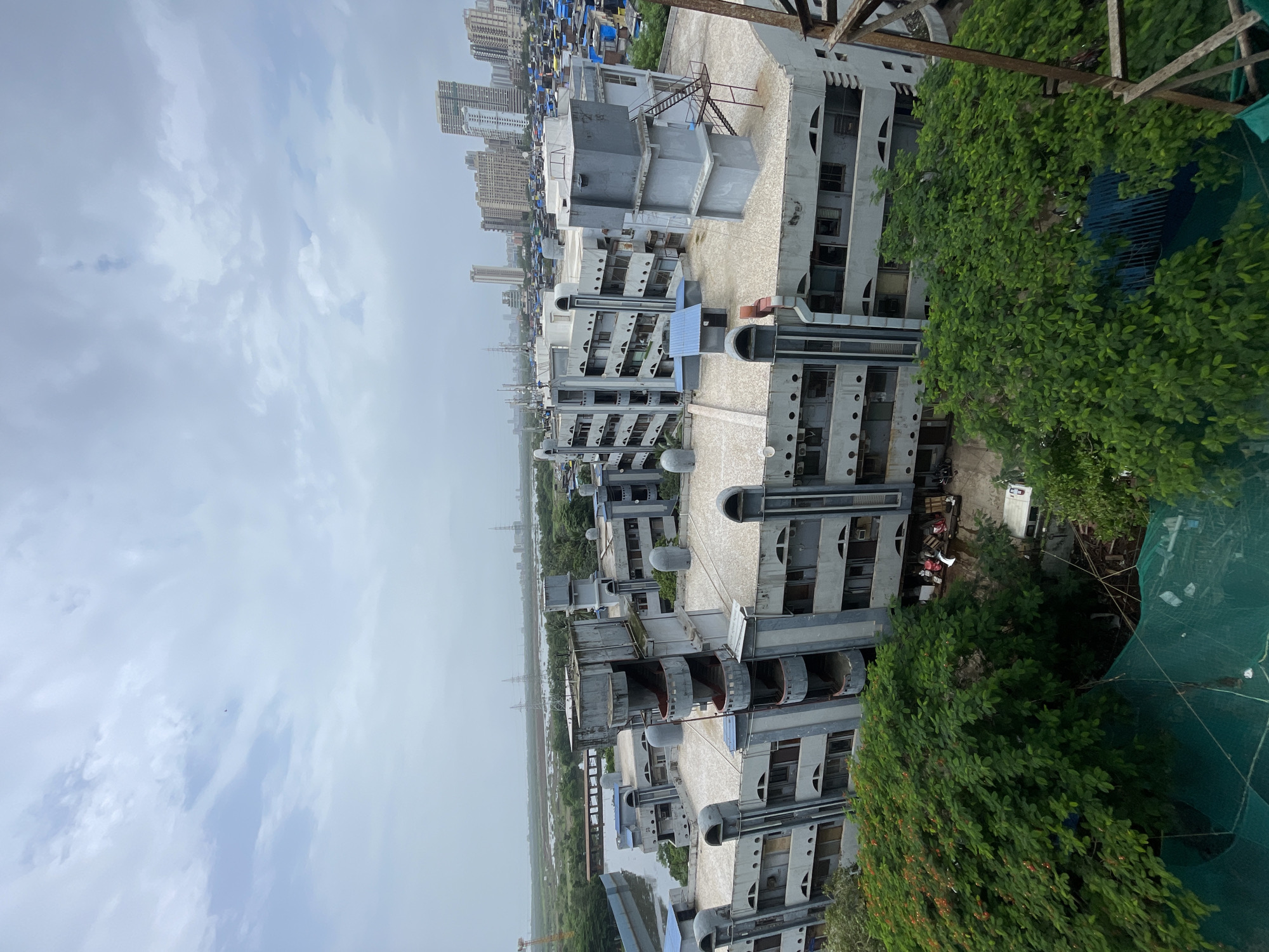 An aerial view captures a cluster of buildings in an urban environment, likely in Mumbai, India. The foreground is dominated by lush green trees, obscuring a portion of the lower buildings. The primary structure in the mid-ground is a large, grey, multi-story building with a distinct, somewhat brutalist architectural style. This building features numerous windows, balconies, and external staircases. Adjacent to it, and further into the background, are more buildings of varying heights and designs, including residential high-rises and densely packed, smaller structures that suggest a slum or informal settlement.
The mid-distance reveals a body of water, possibly a creek or a reclaimed area, with a bridge or walkway spanning across it. In the far distance, the skyline is punctuated by tall skyscrapers, typical of a major metropolitan area. The sky is overcast with clouds, indicating a possibly humid or rainy day. Utility poles and transmission towers are visible in the landscape, suggesting the presence of power infrastructure. The overall scene depicts a contrast between older, institutional-looking architecture, informal settlements, and modern development within a coastal or riverine setting. No people are clearly discernible in the image.