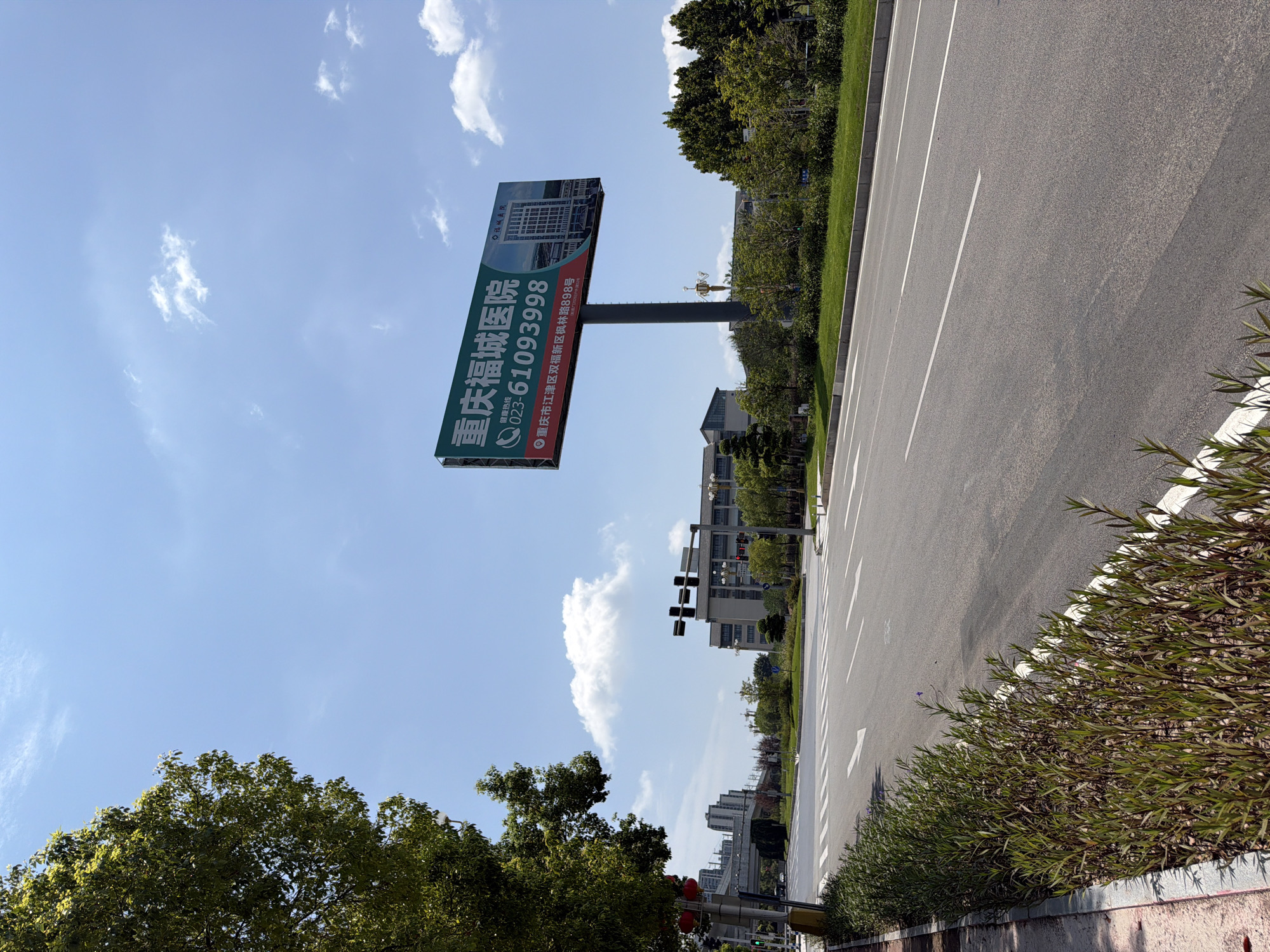 The image shows a wide, empty asphalt road in Jiangjin District, Chongqing, China. A pedestrian crossing with white stripes is visible on the road. On the right side of the road, there is a large green billboard advertising Chongqing Fucheng Hospital. The billboard displays the hospital's name in Chinese characters, a phone number (023-61093998), and its address: No. 898, Xinglin Road, Shuangfu New Area, Jiangjin District, Chongqing. The hospital's building is depicted on the billboard. On the left side of the road, a large tree with green foliage partially obscures the view. In the background, several modern buildings and a bridge can be seen. The sky is blue with scattered white clouds, indicating a clear day. The scene is a typical urban or suburban streetscape, likely captured during daylight hours.