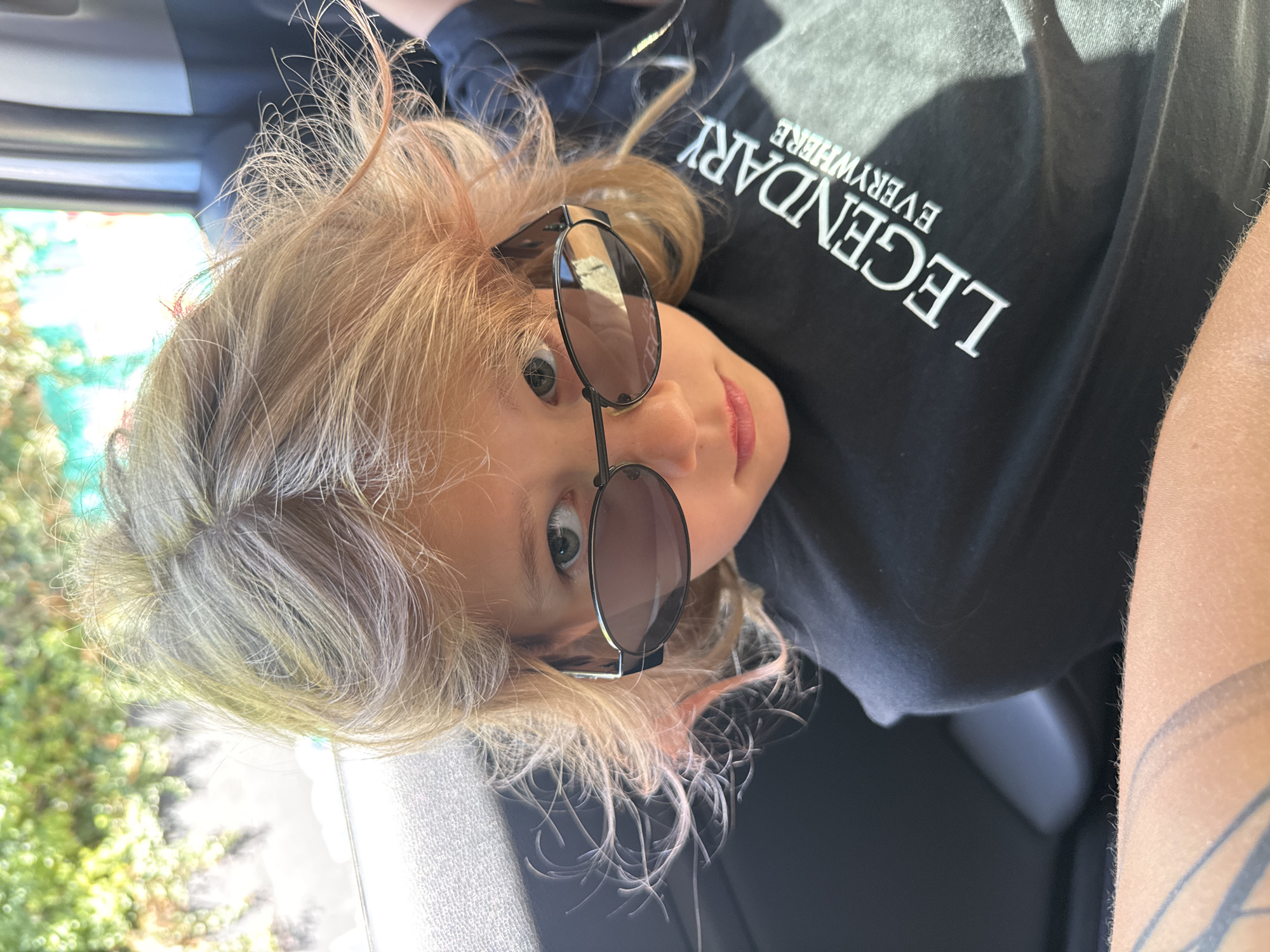 A person with light blonde and gray hair, wearing dark sunglasses and a black t-shirt with the word "LEGENDARY" printed on it, is positioned inside a car. The background features greenery. The location appears to be Kruče - Kërruçi, Montenegro.