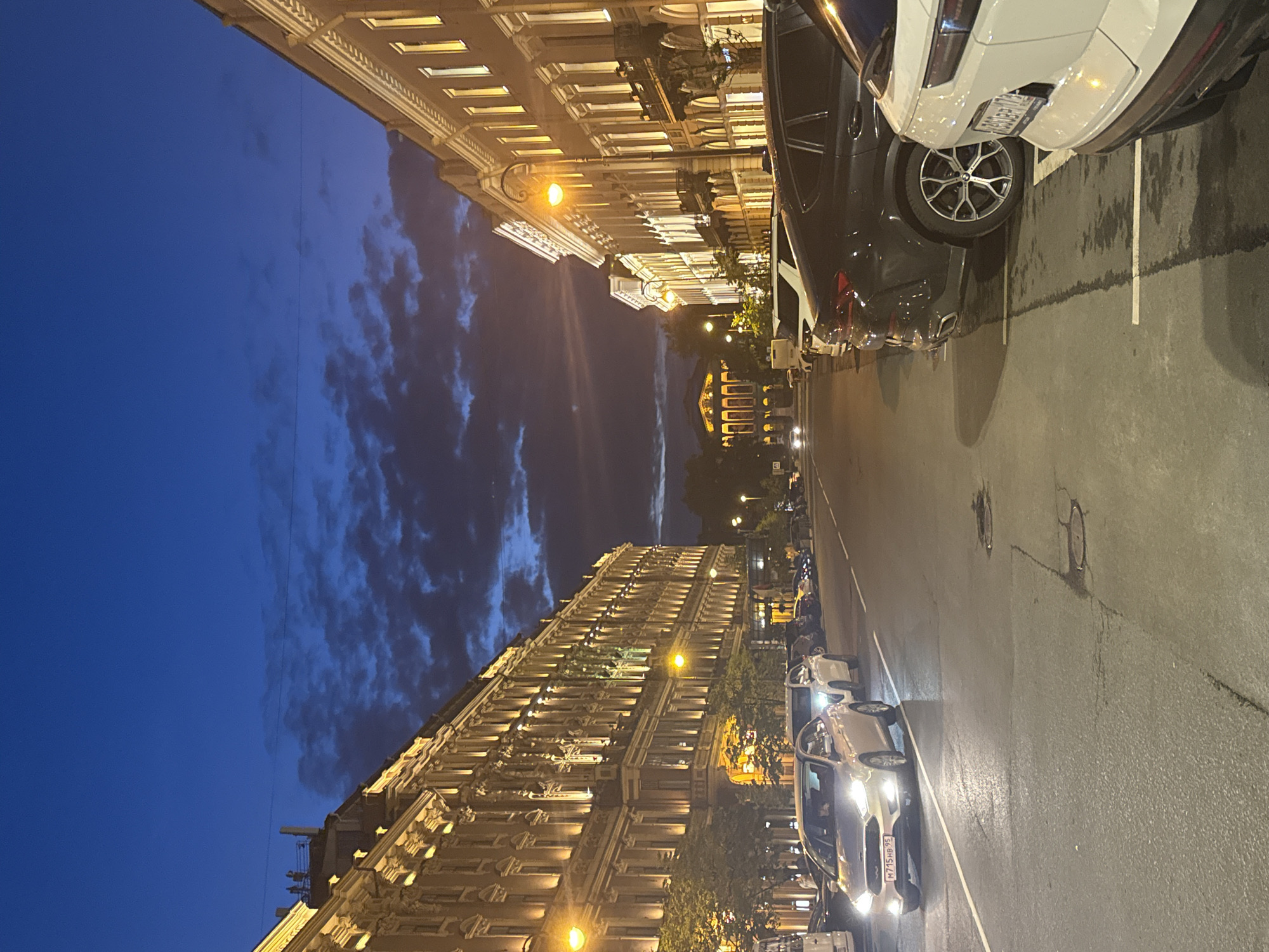 The image depicts a street scene in Saint Petersburg, Russia, at dusk or early night. The sky is a deep blue with scattered, dark clouds. On the left side of the street, a grand, ornate building with many windows and illuminated facade stands tall, its architecture suggesting a historical district. Trees line the sidewalk in front of this building.
On the right side of the street, another building with a similar architectural style and illuminated windows is visible. The street itself is paved and wet, reflecting the lights of the buildings and vehicles. Several cars are parked along the right side of the street, and one car is in motion, driving down the left lane of the street. The parked cars include a white SUV in the foreground on the right, a dark-colored BMW X6, and other vehicles further down the street. The moving car appears to be a silver Ford Focus with its headlights on.
In the distance, at the end of the street, a large, classical-style building with illuminated colonnades and a dome-like structure is visible. Streetlights are intermittently placed along the street. The overall atmosphere is one of urban serenity at the transition from day to night.