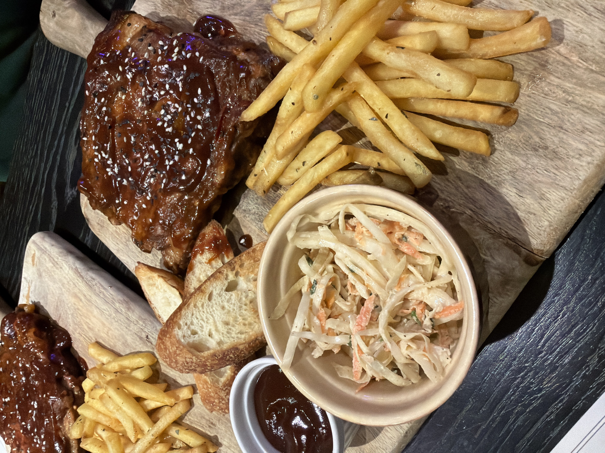 The image presents a close-up, overhead view of a meal served on a wooden cutting board. The main components include a slab of ribs glazed with a dark, possibly barbecue-style sauce and sprinkled with sesame seeds; a generous portion of French fries; a bowl of coleslaw; several slices of bread, and a small ramekin filled with a dark sauce. The food is arranged attractively on the wooden board, which shows signs of use. The board is set on a dark-colored table. This type of cuisine is commonly found in restaurants offering Western or American-style dishes. Given the presence of a dark, possibly barbecue-style sauce on the ribs, the cuisine may be American influenced. Since there is no indication of where the image was taken, it's impossible to tell it's in Tbilisi, Georgia.