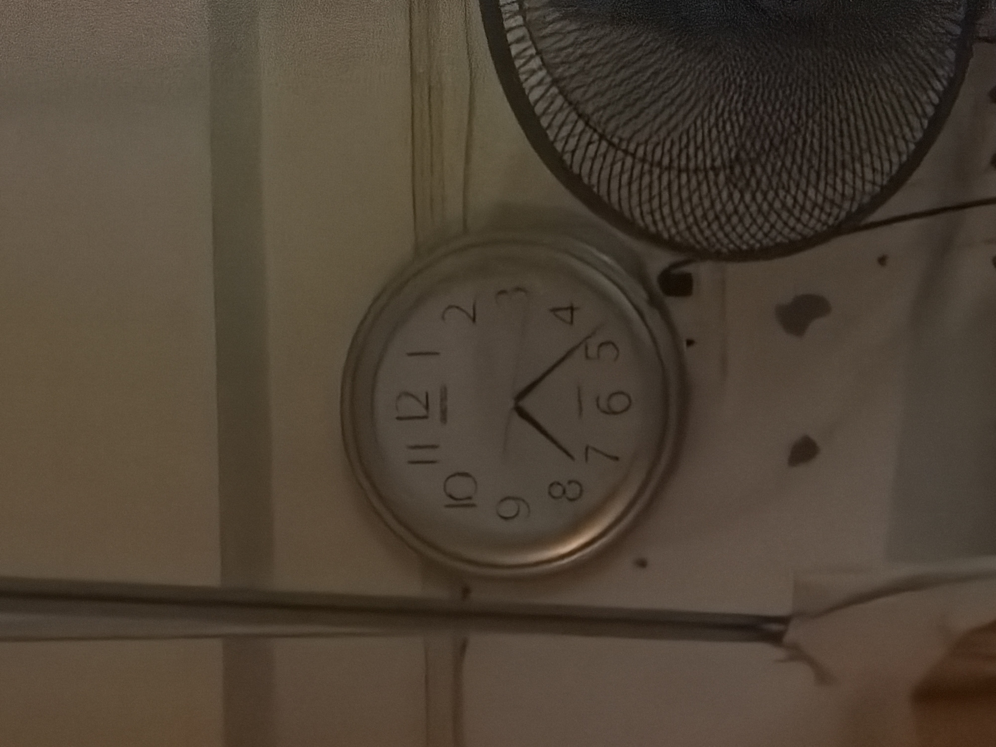 A round clock with black Arabic numerals on a white face is mounted on a light-colored wall. The clock has black hour and minute hands. The hour hand points between the 7 and the 8, and the minute hand points slightly past the 5. To the right of the clock is a black oscillating fan with a metal grate. The background is a textured, light-colored wall with some dark smudges and what appear to be water stains. The scene is indoors, likely in a room in Baan Tai, Thailand.