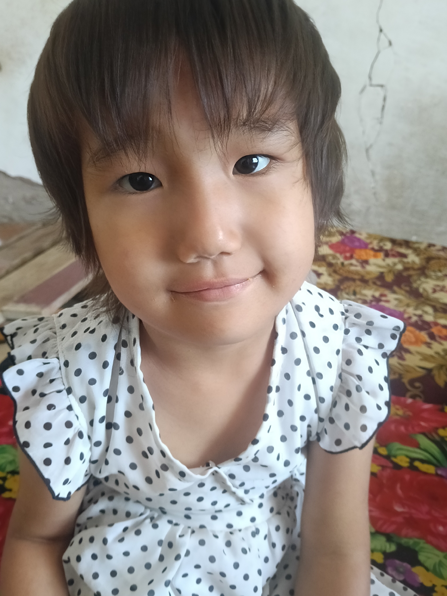 A young girl with dark hair and brown eyes is looking directly at the camera with a slight smile. She is wearing a white dress with black polka dots and ruffles on the sleeves. The background appears to be a room with a patterned bedspread and a plain, cracked wall.