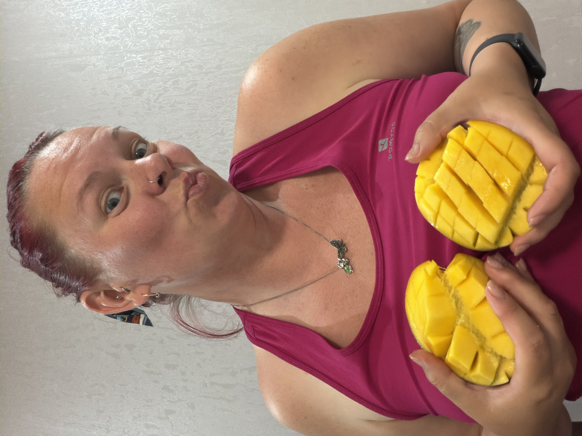 A woman with red hair is holding two peeled and cut mangoes, one in each hand. She is wearing a pink tank top and a silver necklace with a flower pendant. She has a nose ring and earrings. There is a smartwatch on her left wrist, and a tattoo is visible on her left arm. The background is a light-colored wall with a textured pattern. The location is Chaikovsky, Russia.