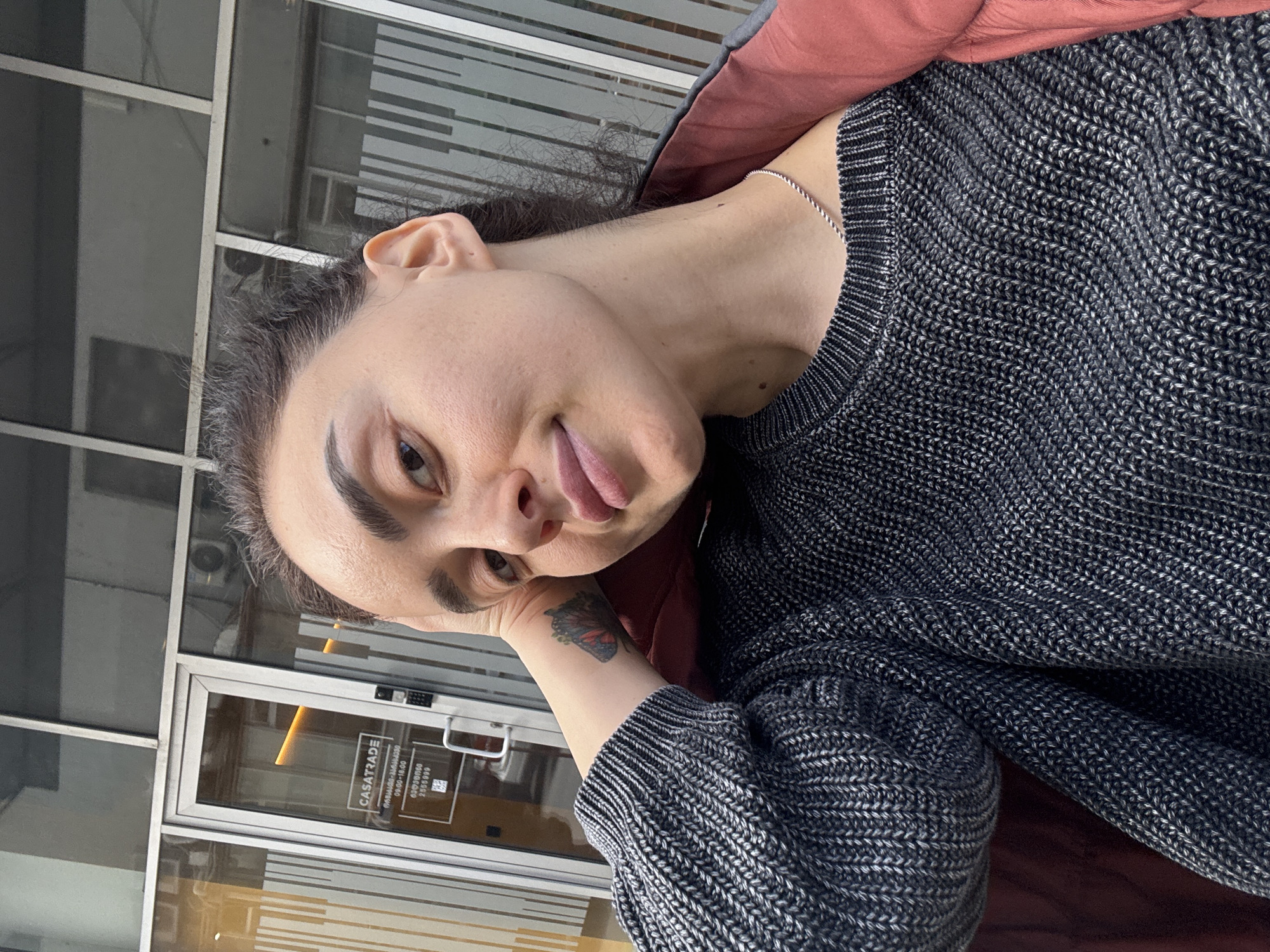A casual portrait features an adult with a light complexion, dark hair pulled back, and prominent eyebrows, set against an indoor backdrop of a glass commercial facade. The person is wearing a textured dark grey/black knitted sweater over a reddish-brown garment, complemented by a thin silver chain necklace. A colorful butterfly tattoo is visible on their left forearm. They look directly at the viewer with a playful expression, slightly sticking out their tongue, while resting their head on their left hand.
The background shows a building's entrance or interior with large glass panels and metal frames, suggesting a shop or office. Reflections are visible on the glass, and through it, parts of an air conditioning unit and other internal structures can be discerned. On the glass door, a white sign with black text is clearly visible. It reads "CASATRADE", "WWW.CASATRADE.GE", and displays business hours "09:00-18:00". Below this, there are numbers "03(0)1/000100" and "255109", possibly phone numbers or identification codes, along with a QR code. The lighting suggests it is daytime. Based on the provided context and the ".GE" domain, the location is Tbilisi, Georgia.