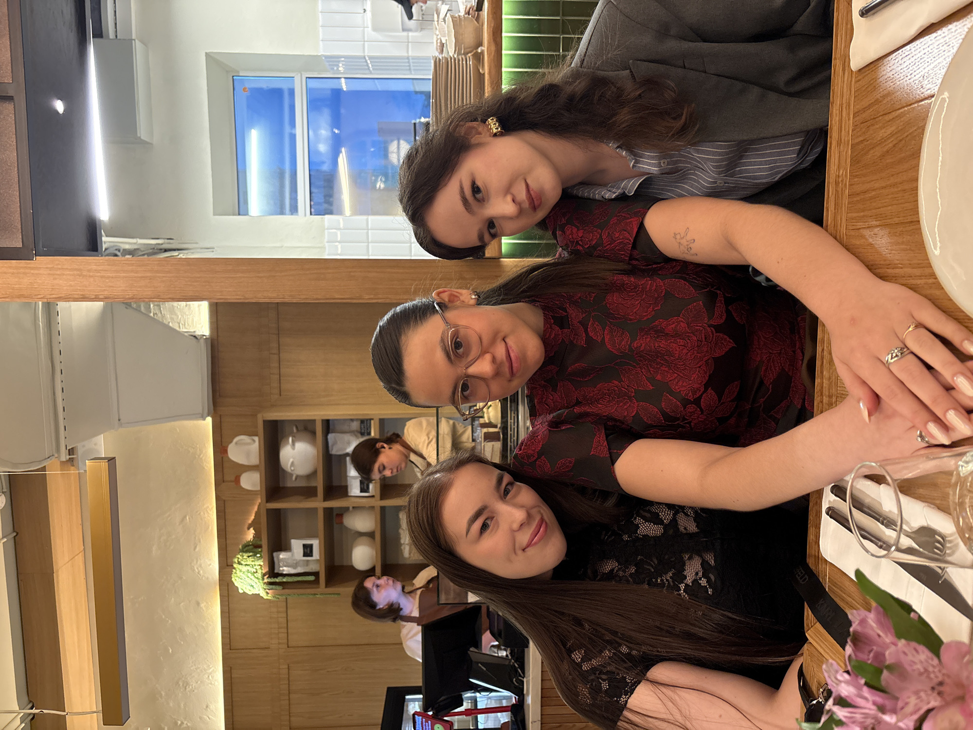 This media file depicts a group of young women gathered around a wooden table in what appears to be a cafe or restaurant. The setting is modern, with clean lines, white tile accents, and warm wooden finishes. There are shelves in the background displaying various white ceramic objects and some greenery.
The primary subjects are three women, positioned in the foreground and mid-ground. The woman closest to the camera, on the right side of the frame, is looking towards the viewer with a smile, her long dark hair cascading down. She is wearing a black lace top. To her left and slightly behind her, another woman with glasses and dark hair is also looking towards the camera, wearing a dark red top with a floral pattern. Her arm is resting on the table, and she is wearing rings on her fingers. A third woman is visible in the background, also looking towards the viewer, wearing a striped shirt and a gray blazer. Her hair is long and dark, and she has noticeable earrings.
The women seem to be engaged in a social gathering, possibly a meal or a casual meetup. The mood appears relaxed and friendly, with the individuals smiling and making eye contact with the camera. The lighting suggests it is daytime, possibly indoors with artificial lighting supplementing natural light from a window visible in the background. The scene is indoors, and the presence of a table setting with cutlery and a glass suggests they are either dining or have just finished. The location context of Perm, Russia, provides a geographical anchor for the scene, though there are no specific visual cues within the image to pinpoint this location beyond the provided context. No discernible text is visible in the image.