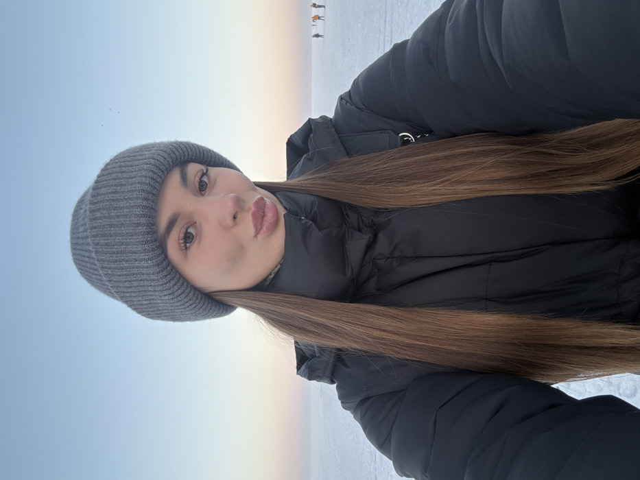 A young woman in a gray beanie and a black puffer jacket poses for a selfie in an outdoor winter setting. She has long, light brown hair and pursed lips. The background shows a pale blue and white expanse, likely snow or ice, with a subtle horizon line tinged with a warm sunset glow. Distant figures are faintly visible on the horizon. The overall impression is of a cold, serene landscape.
