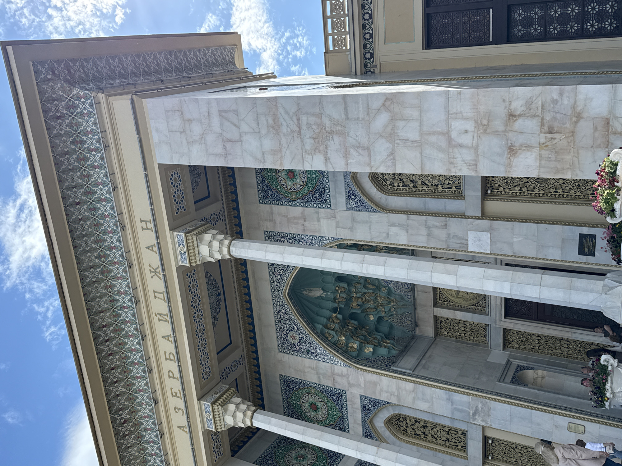 This is a low-angle, wide shot of the exterior of a building, likely a mosque or a cultural center, with intricate architectural details. The building features a facade with marble cladding, ornate tilework in shades of blue, green, and gold, and decorative columns. Above the columns, there's a band of detailed patterned tiles. Some of the tilework includes circular medallions with floral or geometric designs.
The scene is illuminated by natural daylight, suggesting it's daytime. The sky above is blue with scattered white clouds, indicating fair weather. The perspective looks upwards towards the sky and the upper portions of the building, emphasizing its grandeur.
Visible text in Cyrillic letters reads "АЗЕРБАЙДЖАН" (Azerbaijan), suggesting a connection to the country.
Several people are visible in the lower right portion of the frame, appearing to be visitors or attendees. Some are standing near decorative floral arrangements, possibly part of an entrance or celebration. Their presence suggests the location is open to the public and might be hosting an event, or simply receiving visitors. The overall impression is one of cultural significance and architectural beauty.
The building's elaborate design, including decorative archways, carved elements, and intricate tile patterns, points to a location with significant cultural or religious importance, possibly in a city like Moscow, Russia, as indicated by the location context.