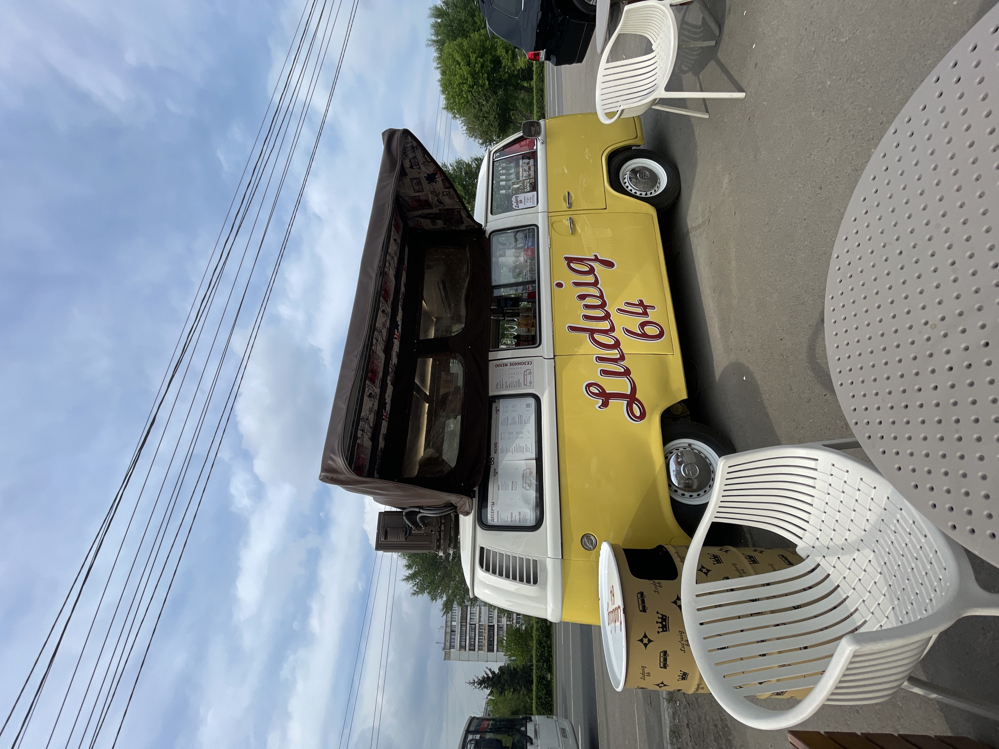 The image depicts a yellow and white vintage Volkswagen van, repurposed as a food truck named "Ludwig 64". The van is parked on an asphalt surface, likely a roadside or parking lot, with a street and a sidewalk visible. In the foreground, there are several white plastic chairs and a circular table with a perforated top, suggesting an outdoor seating area.
To the left of the van, there is a cylindrical object, possibly a trash can or a cooler, decorated with a pattern of crowns, buses, and the "Ludwig 64" logo. A portion of a white bus is visible on the far left of the frame. Behind the food truck, trees and a multi-story apartment building are visible, indicative of an urban or suburban environment. The sky is partly cloudy.
The scene appears to be set in the city of Krasnoyarsk, Russia, as indicated by the presence of a Russian bus and the context of other images from the same location. The food truck is open for business, with menus displayed in the windows and bottles visible inside. There are no people clearly visible in the immediate vicinity of the food truck, though the presence of outdoor seating suggests potential customers. The overall atmosphere is that of a casual, open-air dining or refreshment stop.