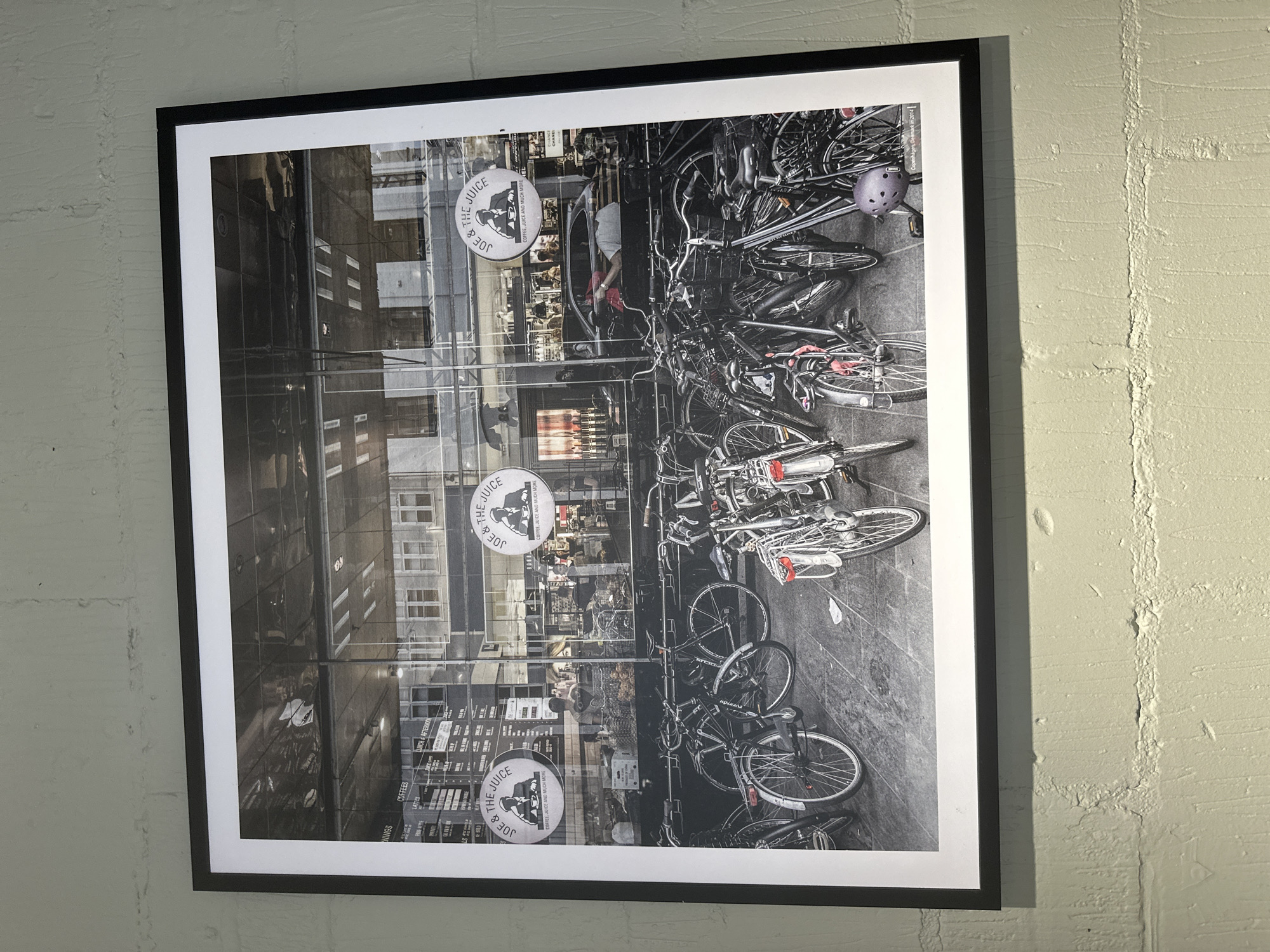 Hanging on a light-colored wall is a framed, black-and-white photograph. The photograph depicts a scene in what appears to be London, United Kingdom. The primary subjects are a row of bicycles parked in front of a "Joe & The Juice" store. The photograph's composition showcases the bustling urban environment, capturing the reflections of the surrounding buildings in the store's glass façade. The photograph appears to be taken from a street-level perspective, and focuses on the bicycles and the store front.