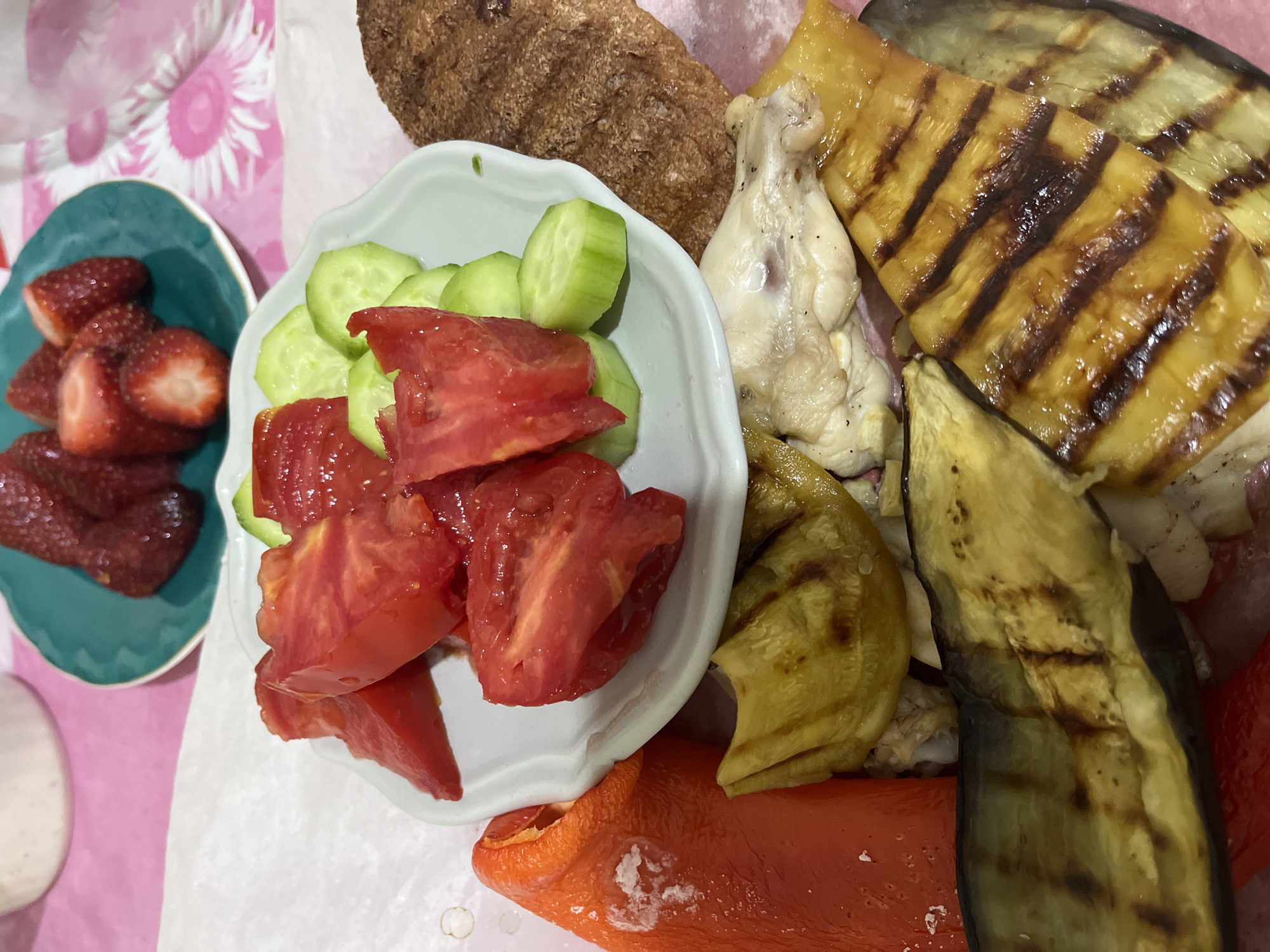 The image shows a spread of food on a table. A turquoise plate holds cut strawberries. Next to it is a white plate with sliced cucumbers and diced tomatoes. Grilled eggplant and peppers, and a piece of bread, are also present. A piece of cooked chicken is visible amongst the grilled vegetables. The background appears to be a tablecloth with a floral pattern. There is no information in the image to identify it as Alanya, Turkey.