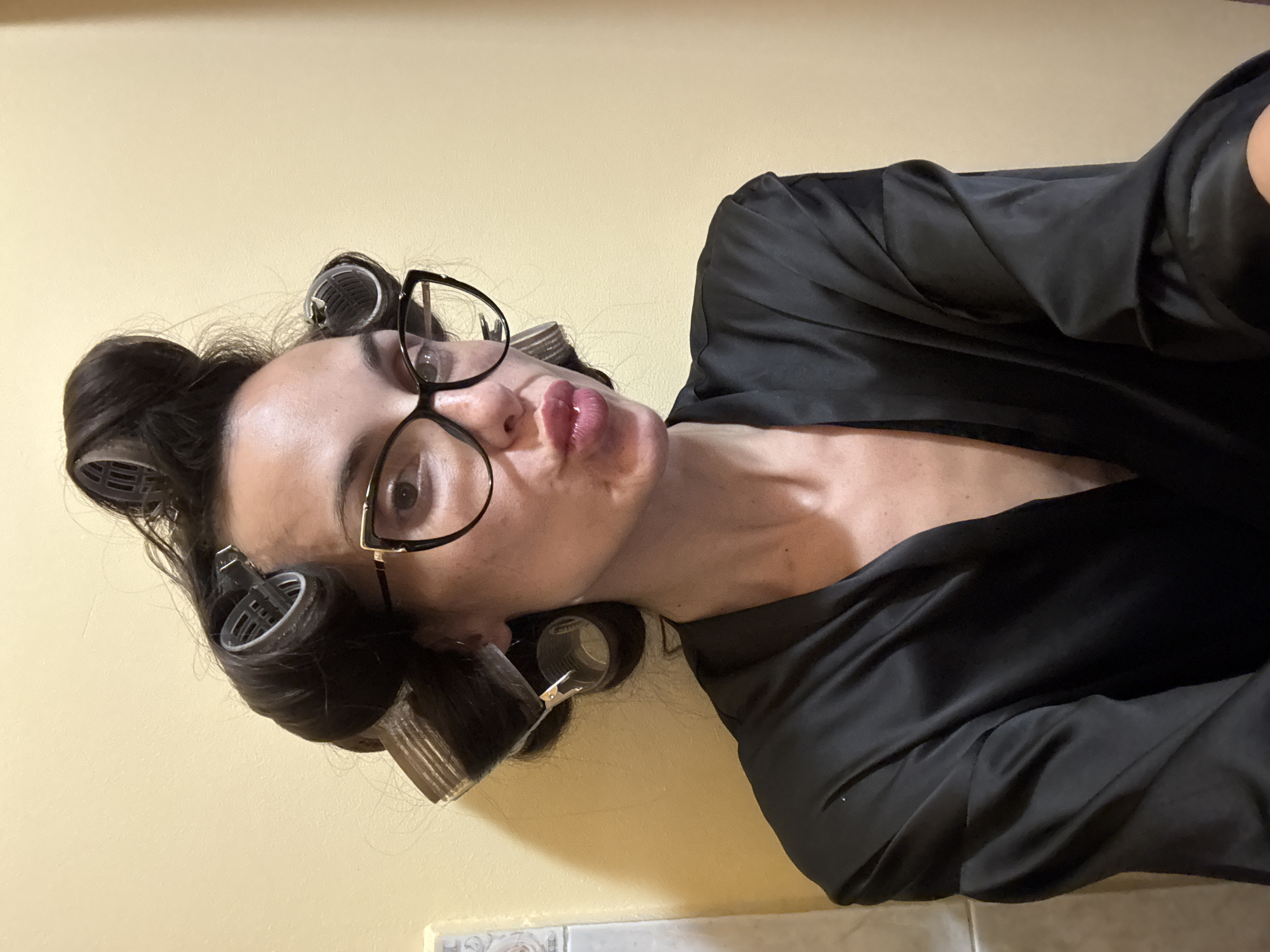 A woman is posing for a photo. She is wearing glasses, a black satin robe, and has hair rollers in her hair. The background is a light yellow wall and a glimpse of a tiled floor. The location is unknown and there is no indication of Pleven, Bulgaria.