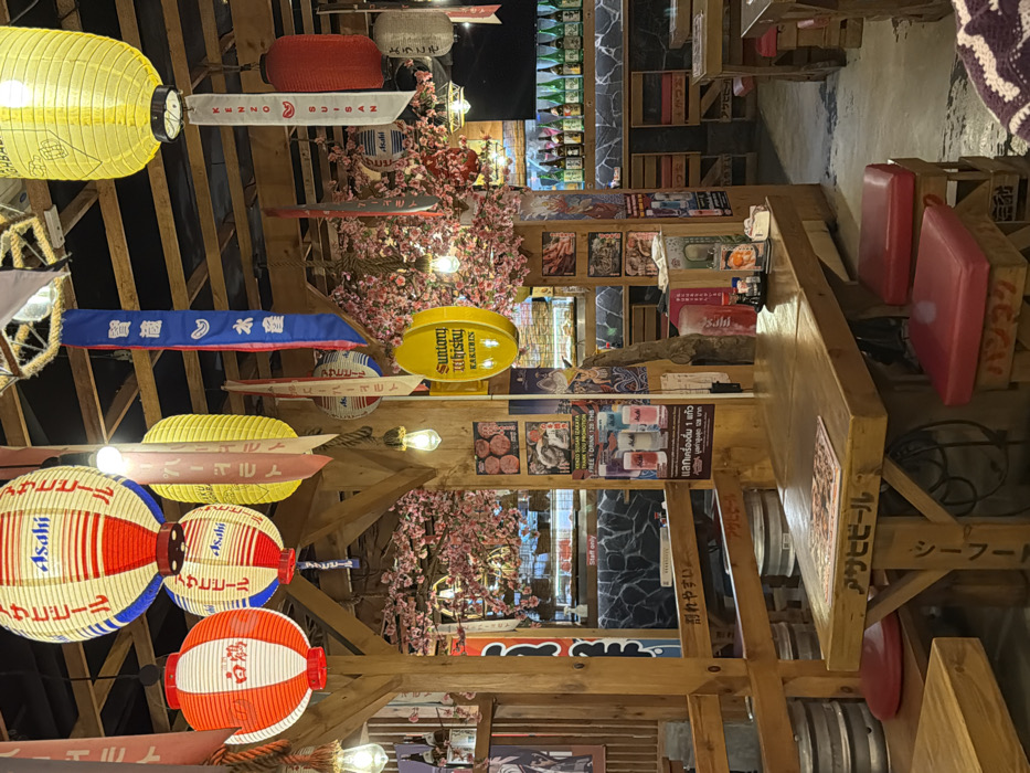 This image depicts the interior of a Japanese-themed restaurant or izakaya located in Bangkok, Thailand. The scene is brightly lit with warm incandescent bulbs and numerous paper and fabric lanterns of various colors (red, white, yellow, blue) suspended from the wooden-beamed ceiling. Several lanterns display "Asahi" branding, while banners and flags with Japanese script, including one for "KENZO SUISAN," also hang throughout the space.
Artificial pink cherry blossoms (sakura) are extensively draped around the wooden structural elements and across the ceiling, creating a festive atmosphere. The furniture consists of robust wooden tables with matching benches, many featuring red cushioned seats. One prominent table in the foreground has "アサヒビール" (Asahi Beer) carved or printed on its side. Metal beer kegs are visible beneath some seating areas.
Vertical wooden pillars are adorned with various posters promoting food items, beverages like Asahi beer and Suntory Whisky Kakubin, and a "KENZO SUISAN" promotion priced in Thai Baht (THB). A bright yellow oval sign for "Suntory Whisky Kakubin" is centrally visible. In the background, behind a counter with a dark, textured stone facade, a display case contains food items. Further back, a bar-like area features shelves displaying numerous bottles, likely sake or spirits. A "Staff only" sign is posted on a counter. The flooring is a light-colored concrete. No individuals are explicitly visible in the frame, though a white crocheted item in the bottom right suggests a partial view of a potential diner or their belonging. The restaurant appears unoccupied or sparsely attended at the time of the photograph.