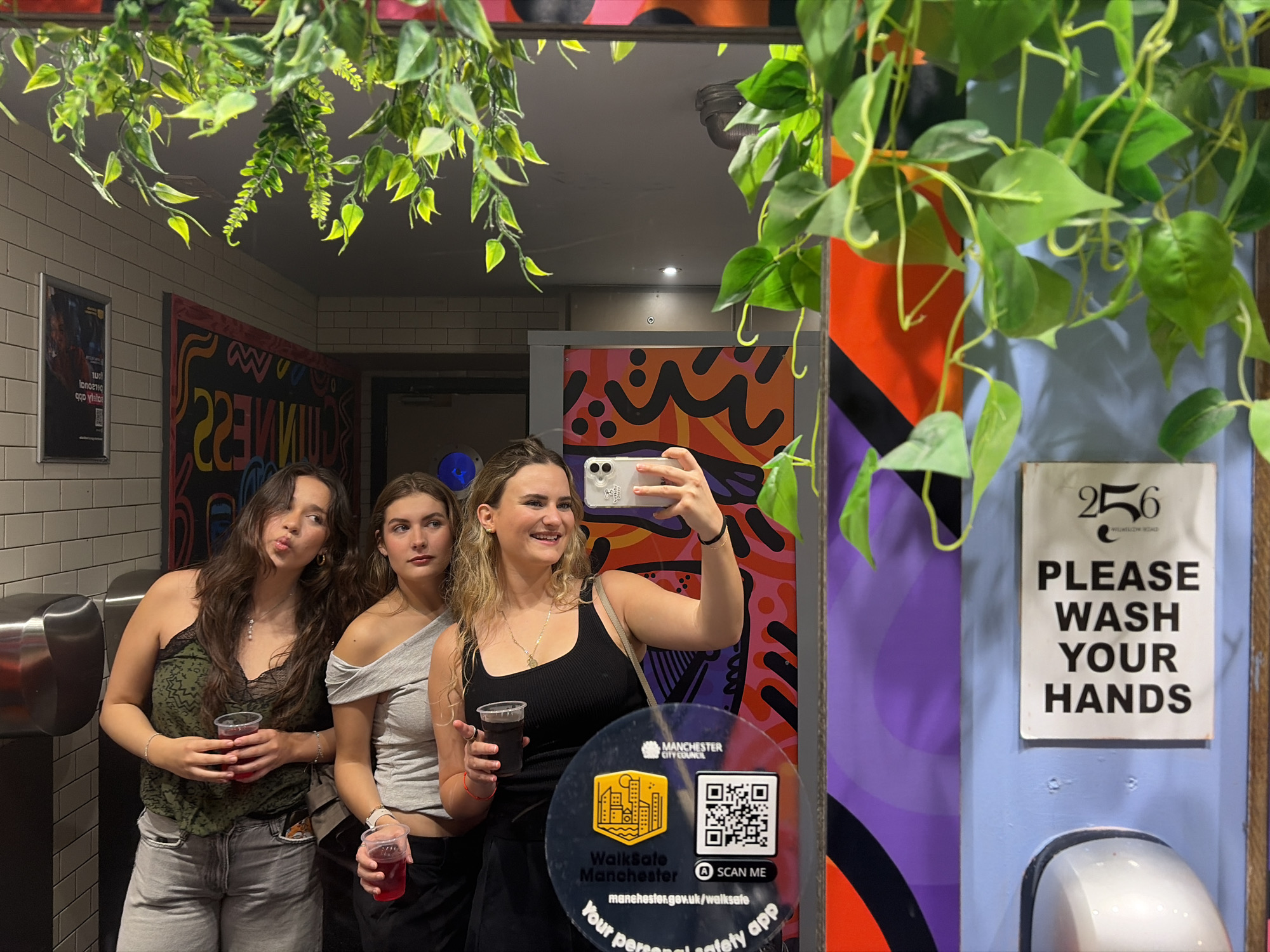 Three young women pose in a bathroom mirror, each holding a red beverage. The woman on the right holds a cell phone, presumably taking the photo. The wall behind them is adorned with colorful abstract designs. A sign reading "PLEASE WASH YOUR HANDS" is visible. A logo for "WalkSafe Manchester" is affixed to the mirror. The OCR results suggest that the location is in Manchester.