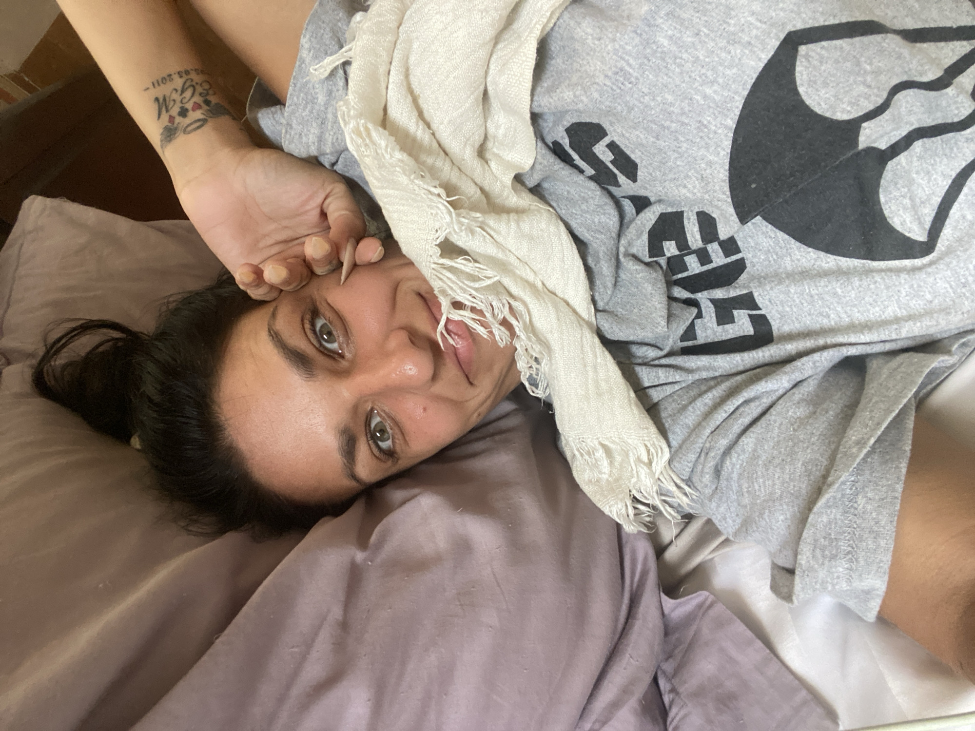 A woman with dark hair and brown eyes, wearing a grey t-shirt and shorts, lies in bed. She has a tattoo on her left forearm. A beige scarf is draped over her. She is looking at the camera and smiling, with her left hand resting on her cheek. The background consists of pillows and bedding. The scene appears to be indoors, in a bedroom. There is no indication of the city of Mũi Né, Vietnam.