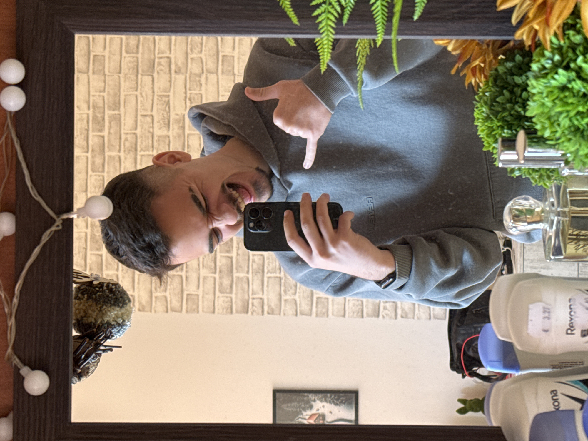 A man is reflected in a mirror with a brown frame. He is holding a black smartphone and pointing down with both hands, while smiling and making a playful, slightly exaggerated expression. The man is wearing a gray hooded sweatshirt. The background visible in the mirror includes a light-colored brick-patterned wall. The area around the mirror is decorated with fairy lights, a decorative floral arrangement, toiletries (including Rexona deodorant), and a small framed picture on the wall. There is no verifiable information to locate this scene in Straldzha, Bulgaria.