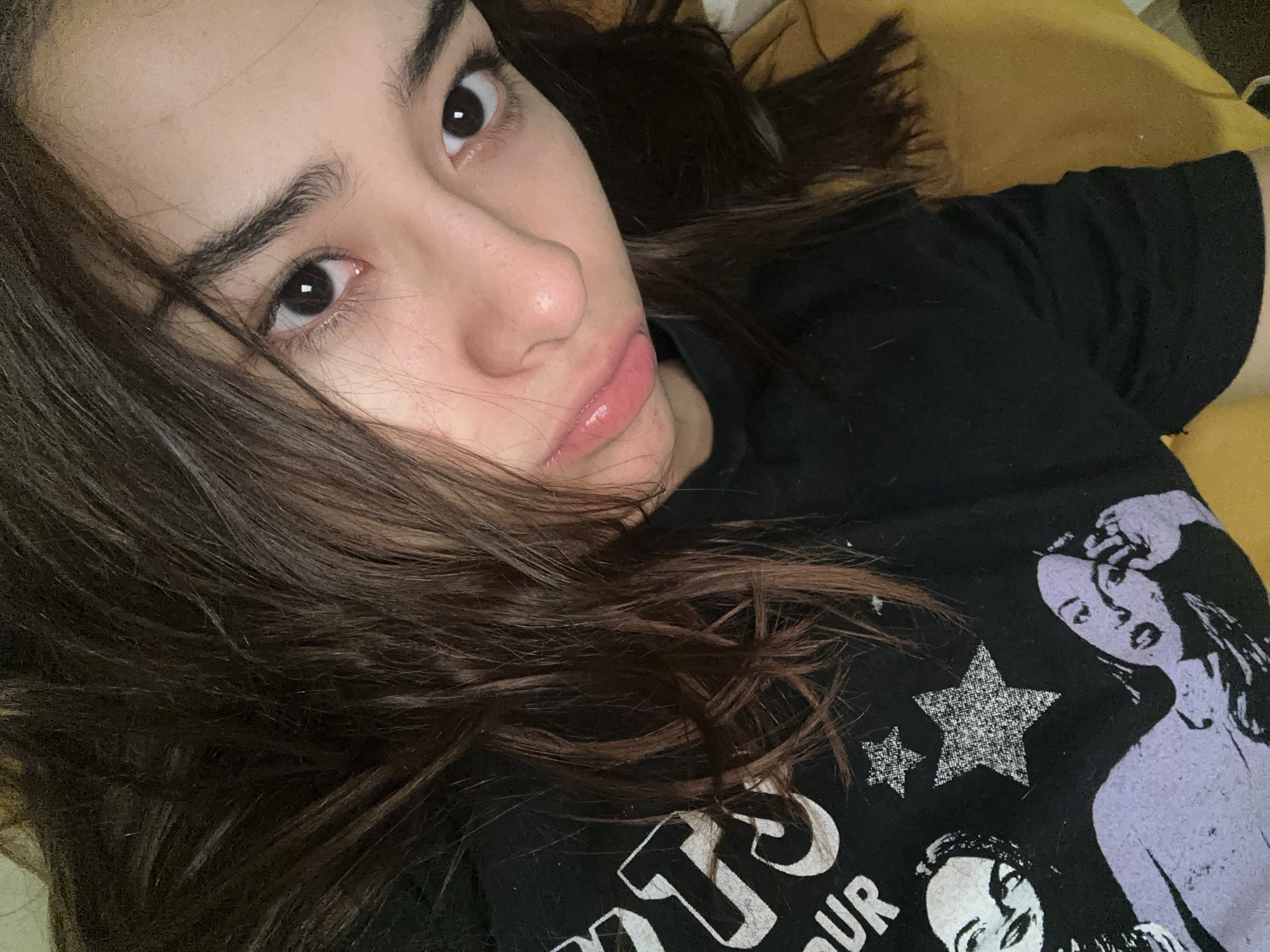A young woman with dark hair and brown eyes lies on a yellow surface, possibly a pillow or cushion. She is wearing a black t-shirt with a design featuring stars and graphic figures. The woman's face is angled towards the camera, showing her features clearly, including her eyebrows, nose, and lips. The environment is indoors, suggested by the presence of the pillow or cushion. There is no indication of the city of Puebla, Mexico in the image.