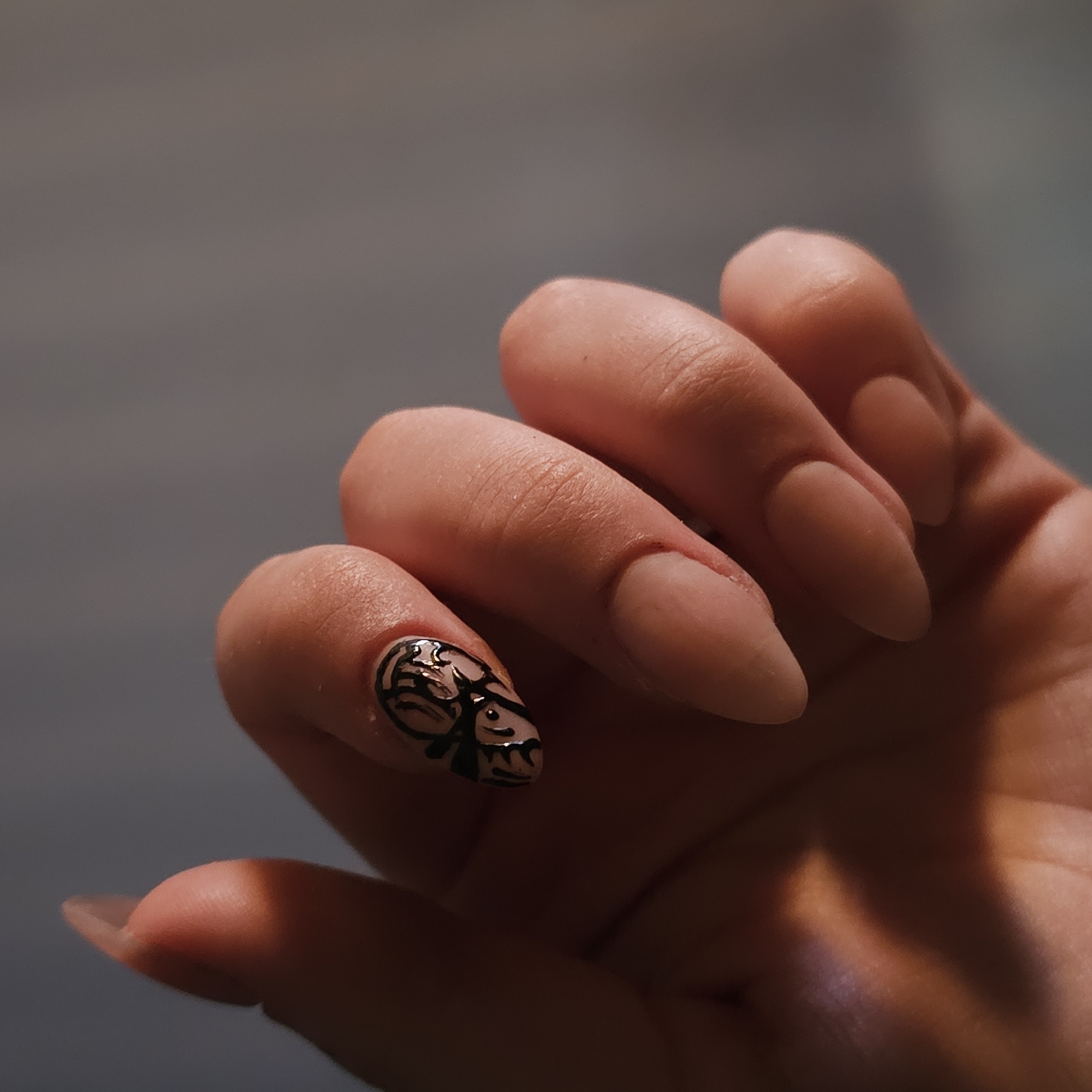The image shows a close-up of a person's hand with almond-shaped fingernails. One fingernail, the index finger, has black tribal-like nail art. The background is a muted gray, suggesting an indoor setting. There is no discernible location in Nizhny Novgorod, Russia.