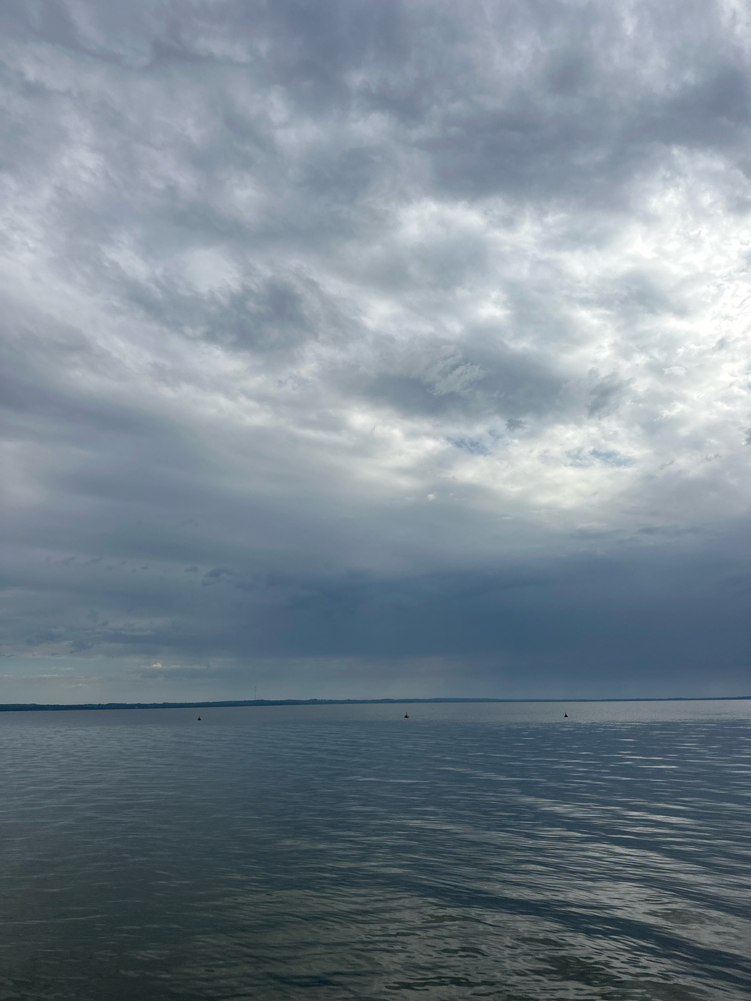 The image depicts a vast expanse of calm water, likely a lake or a large river, under a dramatic, cloudy sky. The water's surface is subtly rippled, reflecting the muted tones of the overcast heavens. A thin, dark line of distant land, possibly with some vegetation, marks the horizon. Three small, red buoys are sparsely distributed across the water, indicating a marked channel or area. The overall mood is somber and expansive, suggesting a tranquil yet potentially imposing natural environment. The location is specified as Нарацкі сельскі Савет, Belarus, which is known for its natural landscapes, including lakes and forests. This scene is consistent with the geography of the region. There are no people, animals, or specific structures visible in the frame, other than the buoys.