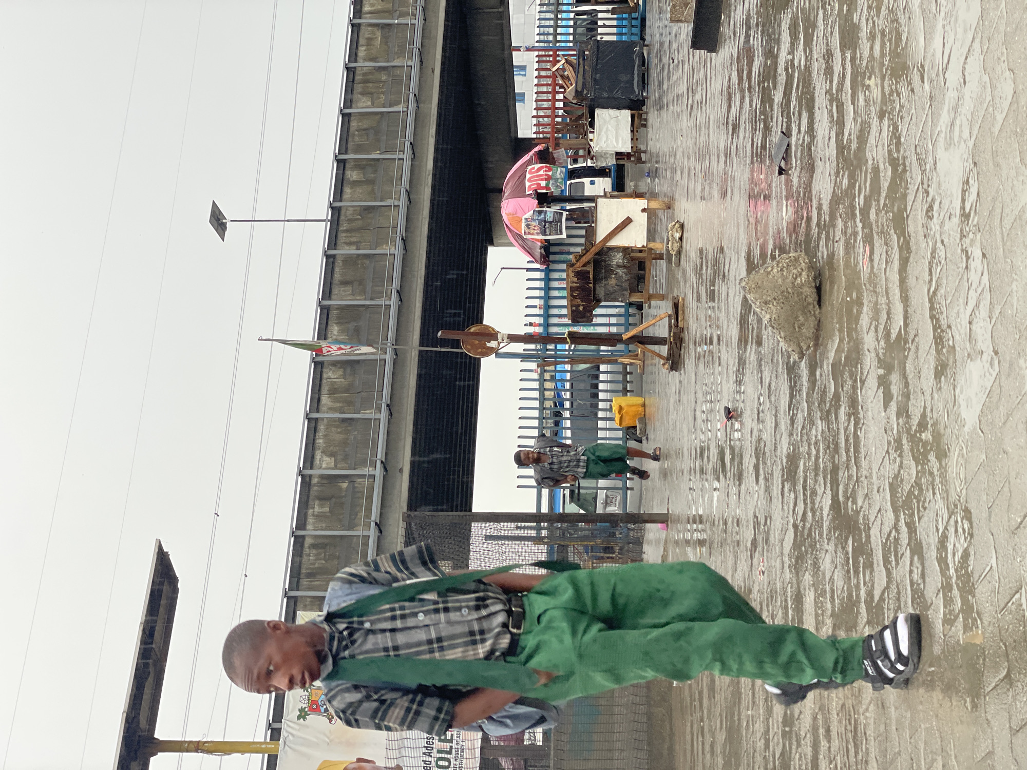 The image shows a street scene in Lagos, Nigeria on a wet day. A man in a plaid shirt and green trousers walks along a paved surface that is covered with water. Behind him is an elevated roadway. There are several stalls or vendors set up along the roadside. Another man is visible sitting by a blue fence. The street is wet and reflects the cloudy sky. There is a flag and light post visible.