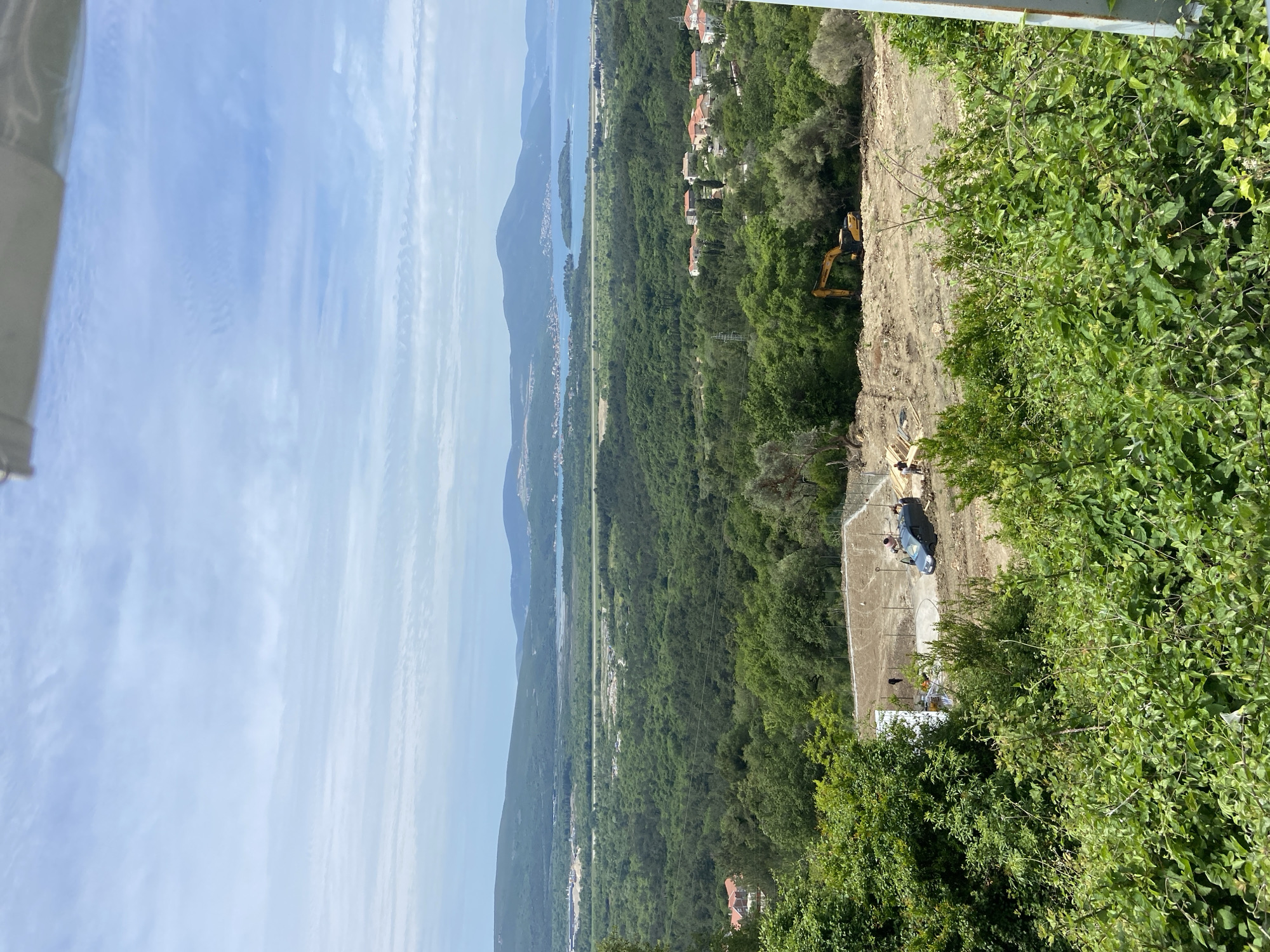 The image depicts a view from an elevated position in Kavač, Montenegro. In the distance, mountains and a body of water are visible under a sky with scattered clouds. In the middle ground, dense green vegetation covers the land. A dirt construction site is present, featuring an excavator and some workers. A concrete structure is also visible in the construction area. Green foliage is prominent in the foreground.