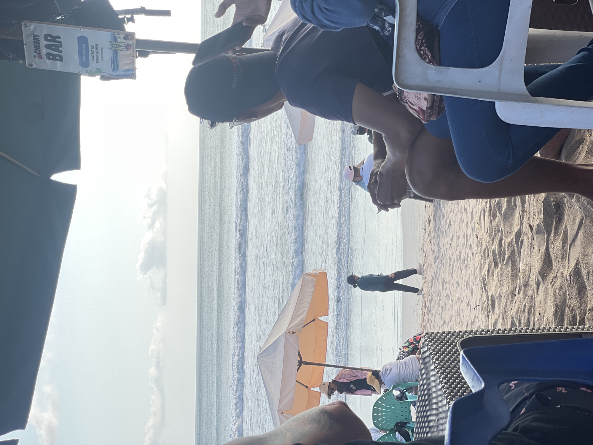 In Kuta, Indonesia, a beach scene unfolds under a canopy, with the ocean and breaking waves visible in the background. In the foreground, several people are seated on chairs, facing the sea. A man in a dark t-shirt and a baseball cap is in the center, holding a phone. To his right, a person wearing blue jeans is seated. To the left, a person in a light-colored top and a straw hat is visible. A beach umbrella is set up on the sand, and further back, a few individuals are walking along the shoreline. A sign for "CHERRY BAR" hangs overhead. The sand is light brown, and the water appears calm with moderate waves.